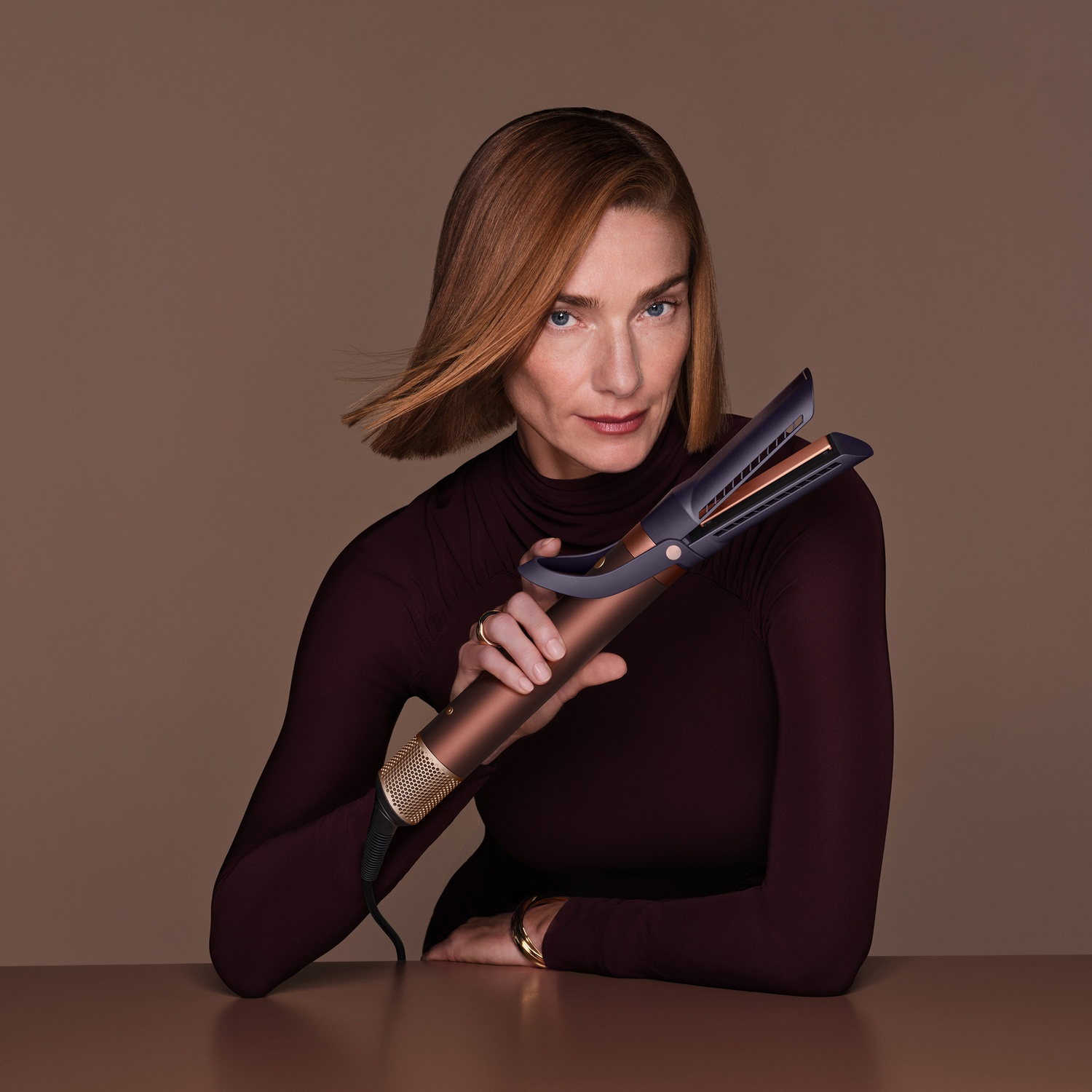 Una donna con i capelli rossi posa su sfondo marrone reggendo un Dyson.