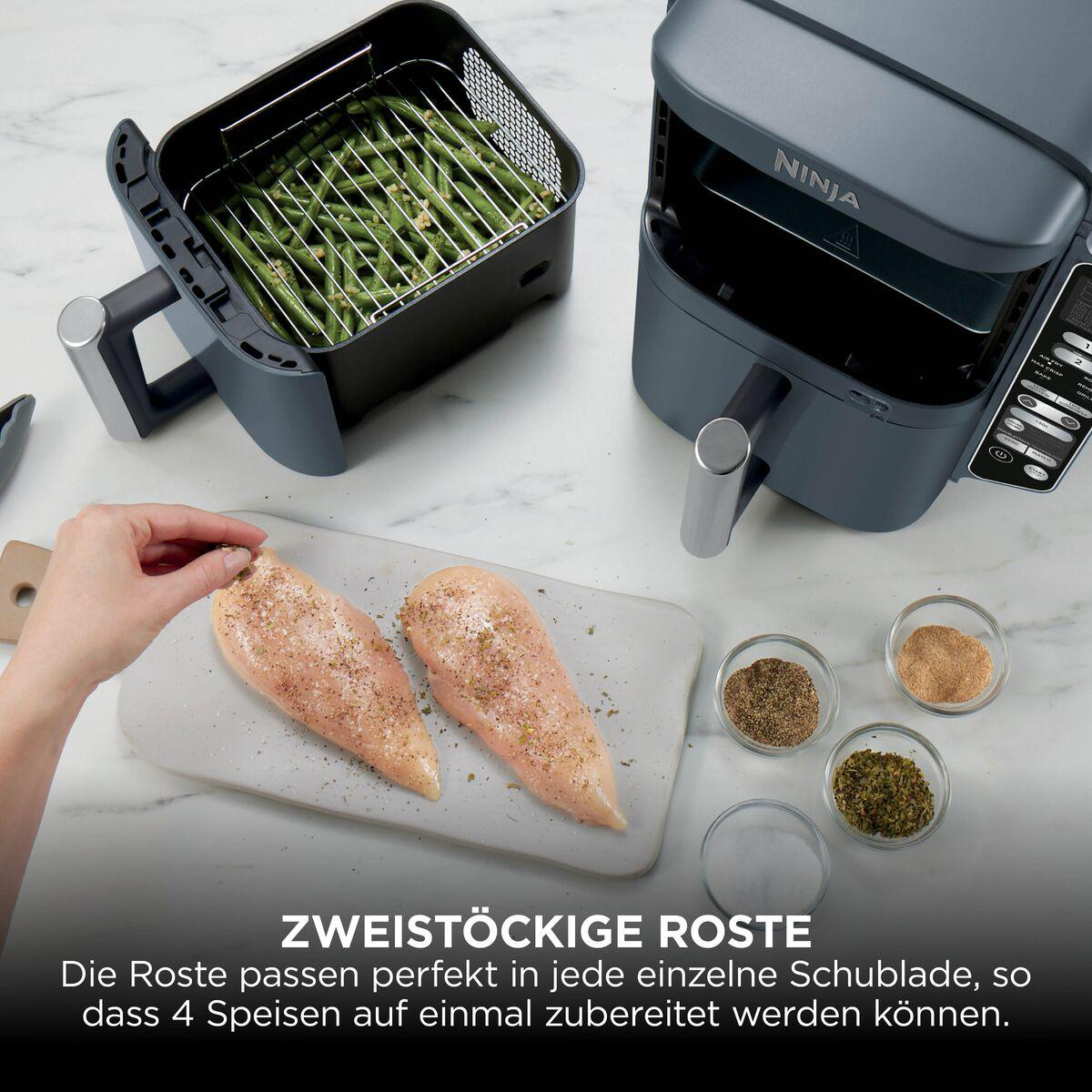 Eine Ninja-Heißluftfritteuse mit grünen Bohnen und rohen Hähnchenbrüsten mit Gewürzen auf einer hellen Arbeitsplatte.
