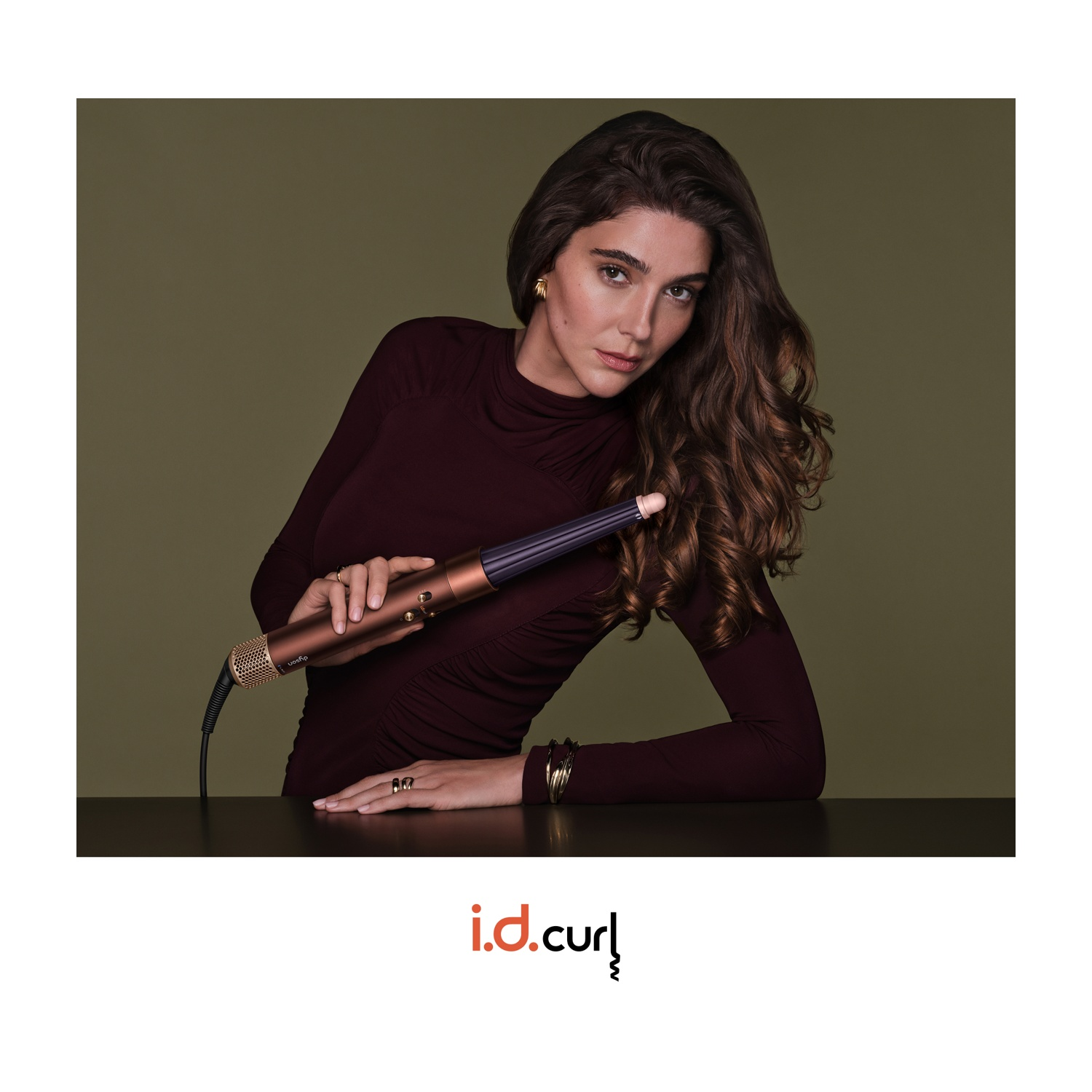 Una donna con un abito bordeaux tiene un ferro arricciacapelli su sfondo verde oliva.