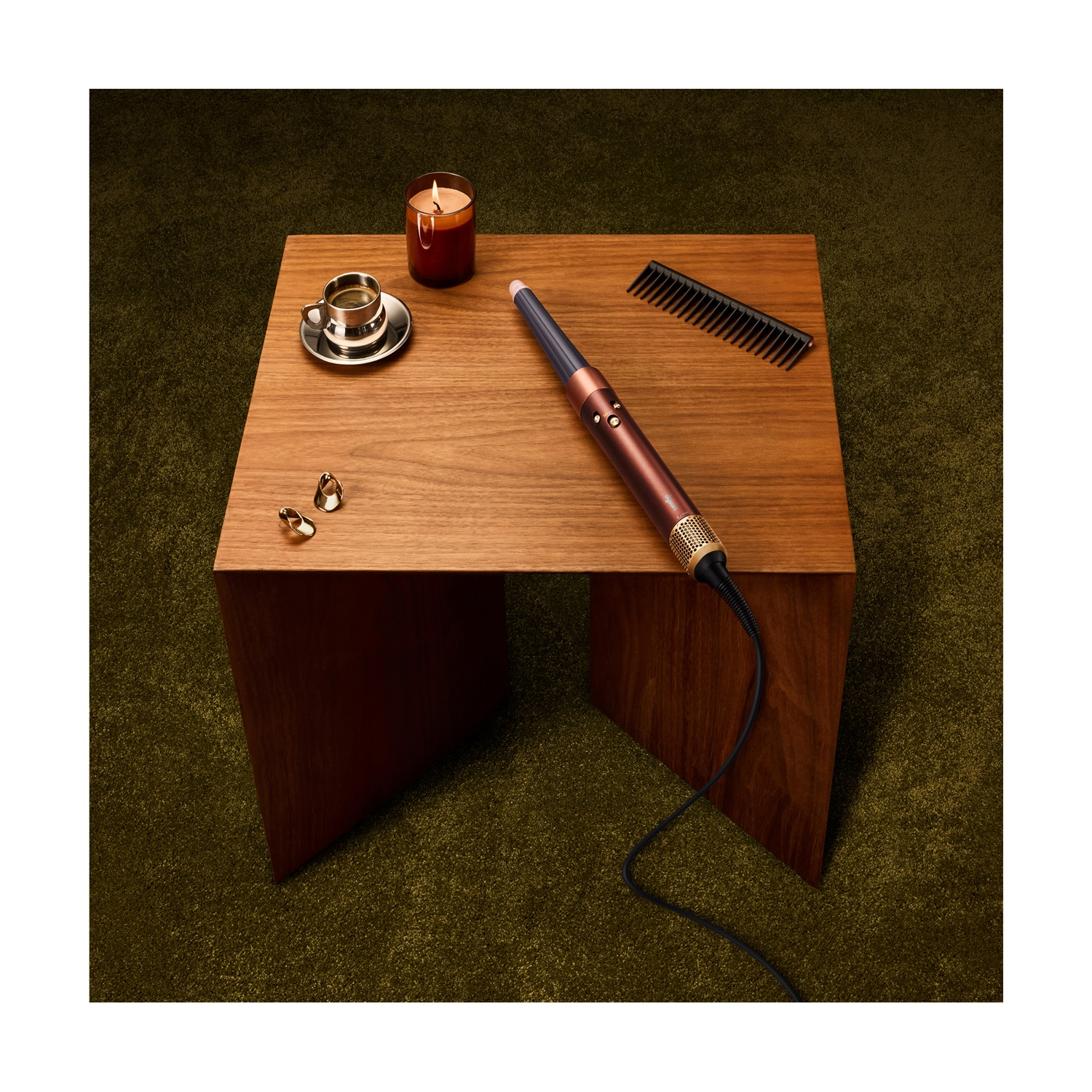 Uno styler, tazza di caffè, candela, pettine e anelli su un tavolo di legno su un tappeto verde.