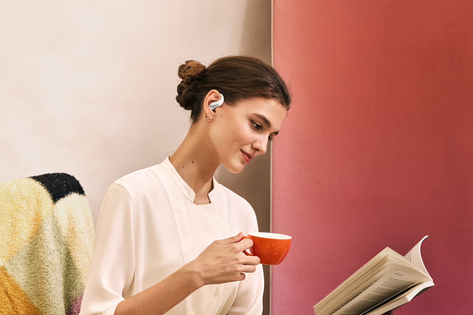 Eine Frau in weißer Bluse liest ein Buch, hält eine rote Tasse und trägt weiße Ohrhörer.
