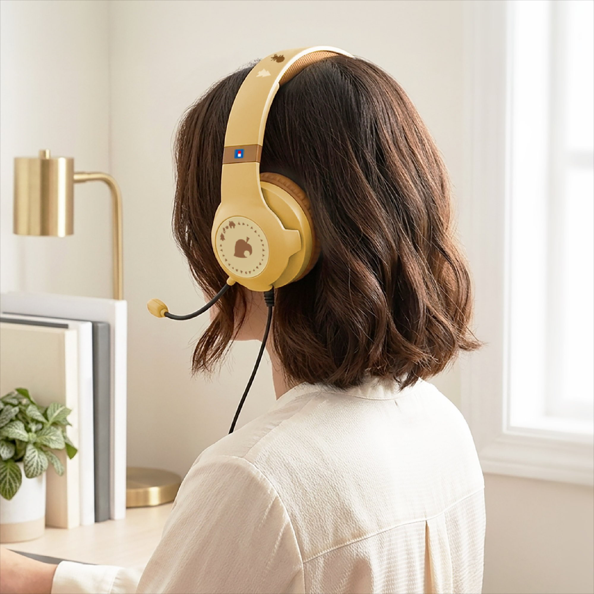 Una donna con i capelli castani indossa cuffie beige con microfono, seduta a una scrivania con libri e lampada.