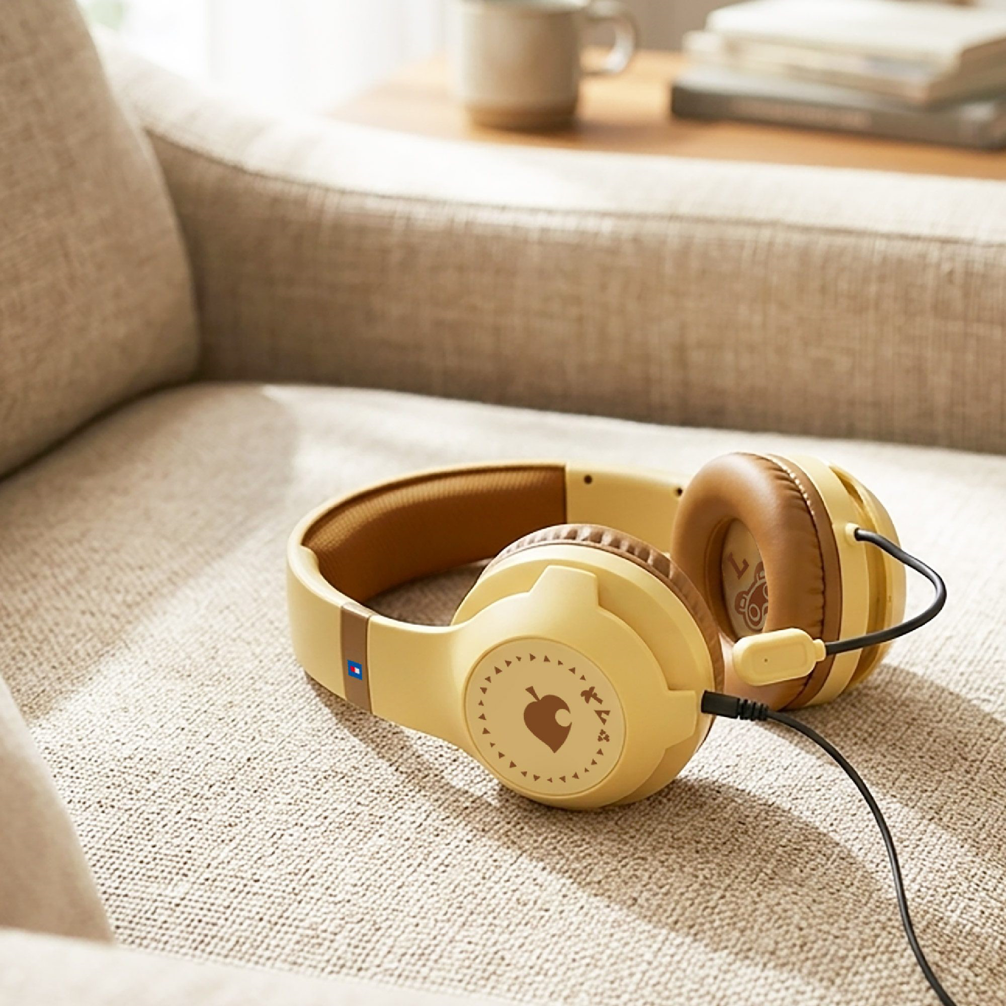 Cuffie beige con dettagli marroni su un divano chiaro, con un tavolo e libri sullo sfondo.