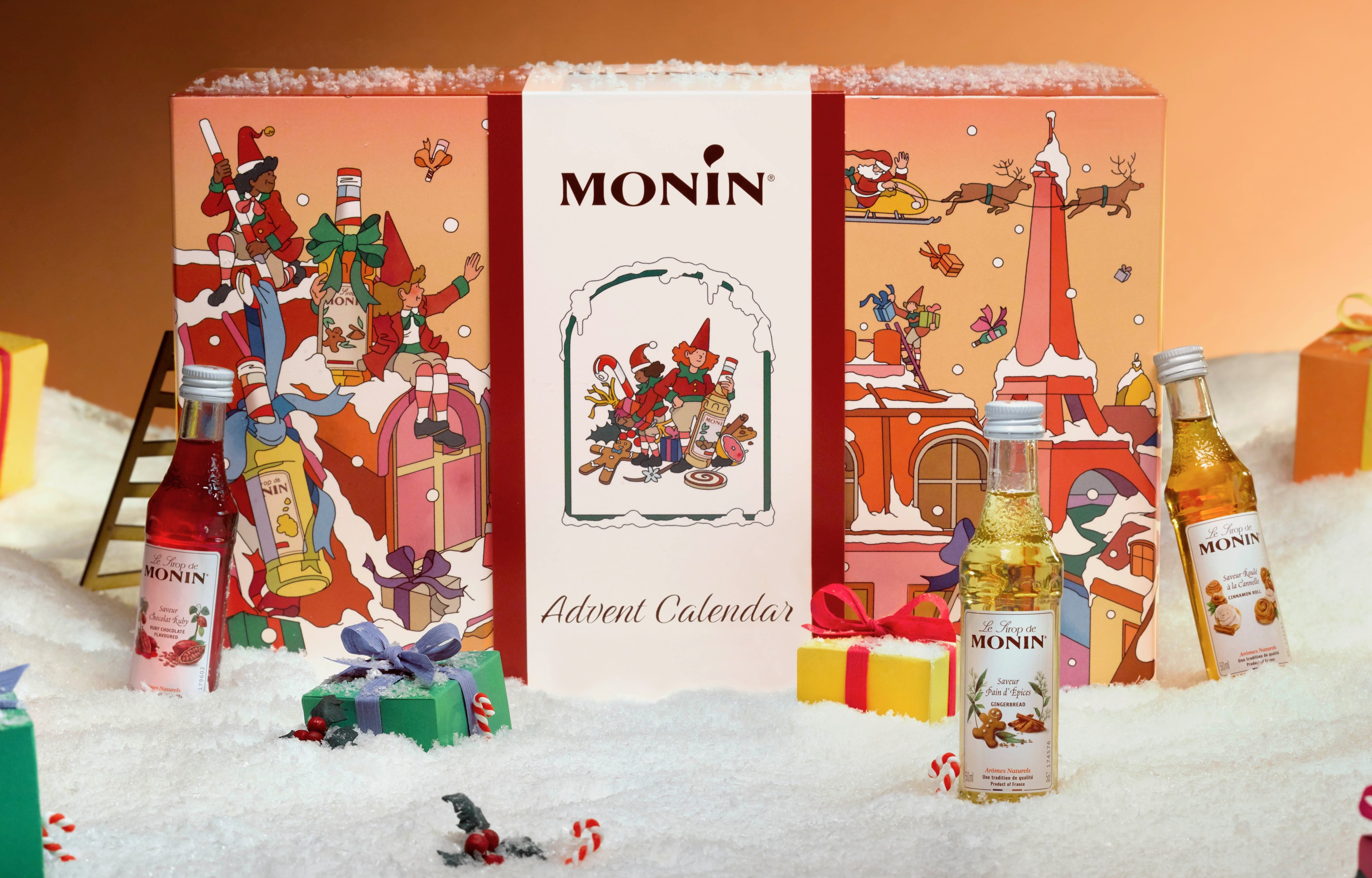 Ein Monin Adventskalender mit Weihnachtsszenen im Schnee und drei kleinen Sirupflaschen davor.
