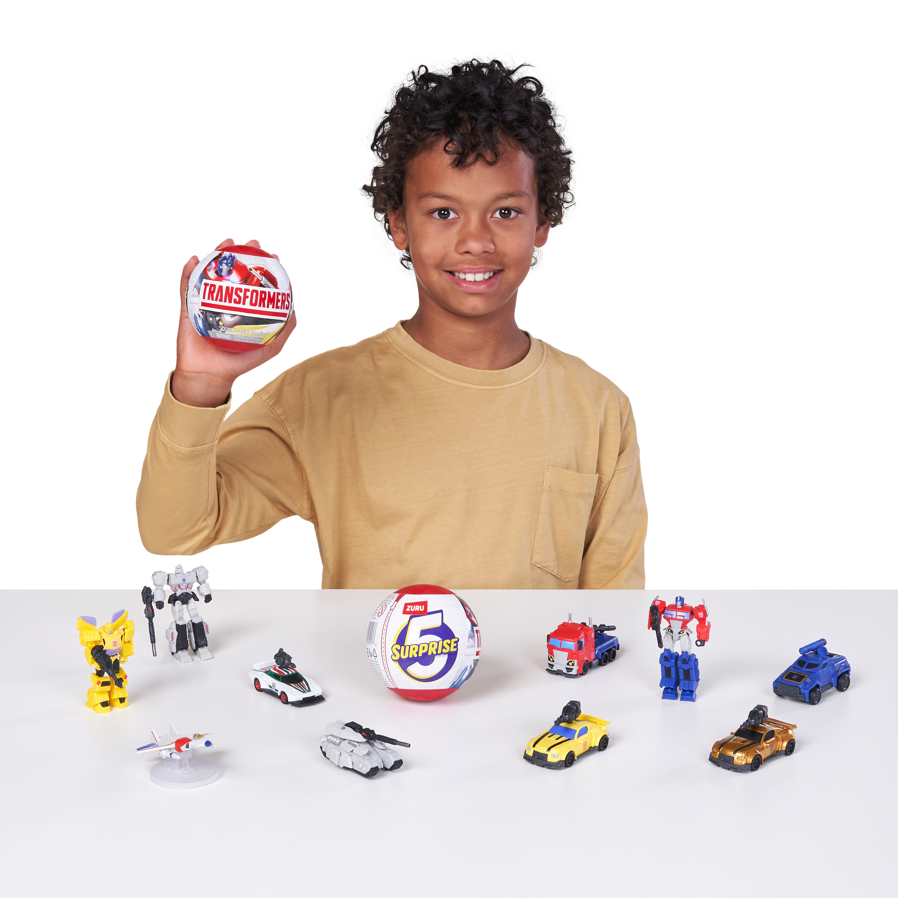 Un niño sonriente con camisa amarilla sostiene una bola de Transformers rodeado de juguetes.