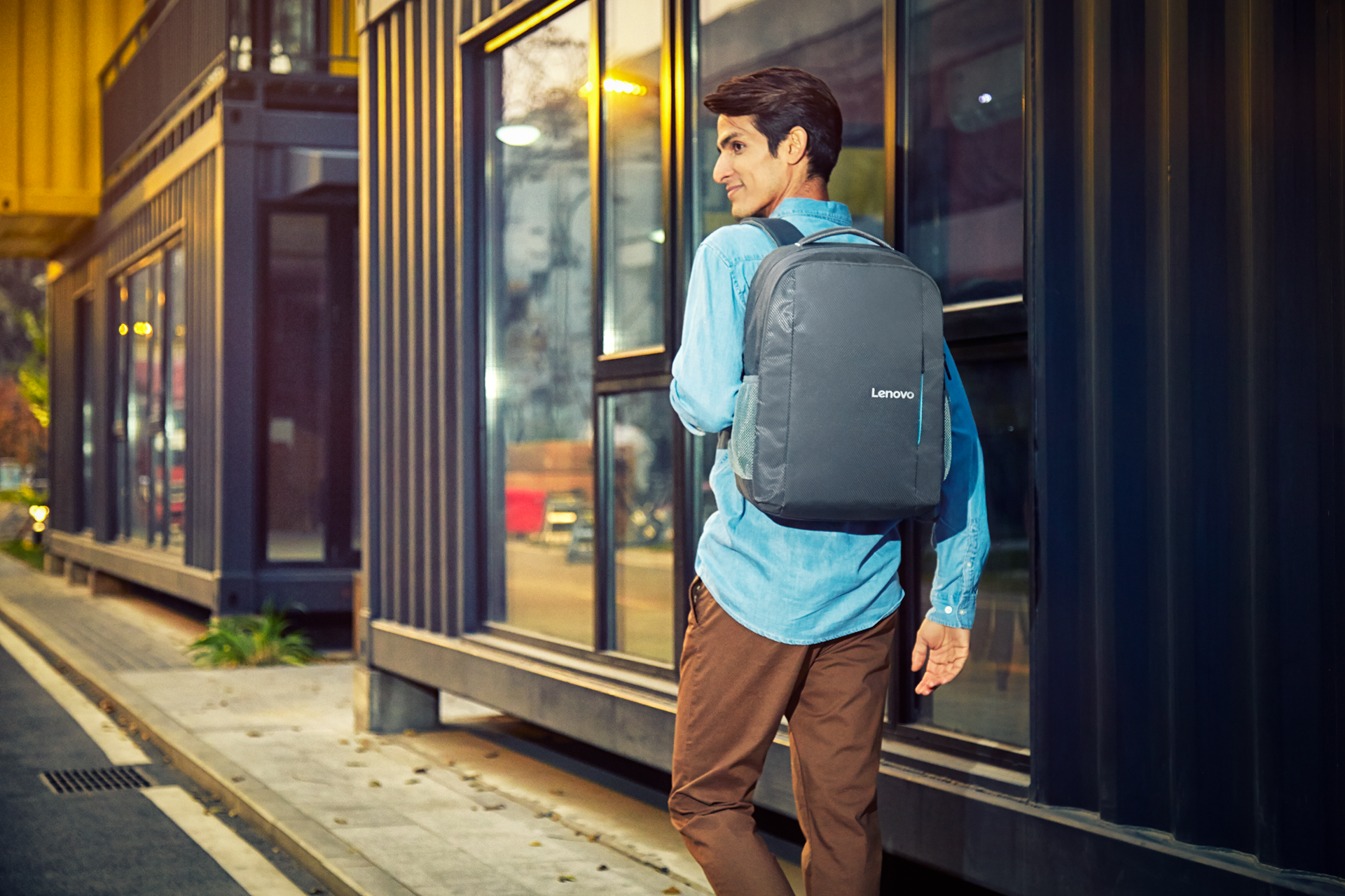 Un joven con camisa azul y mochila gris de Lenovo camina junto a un edificio moderno.