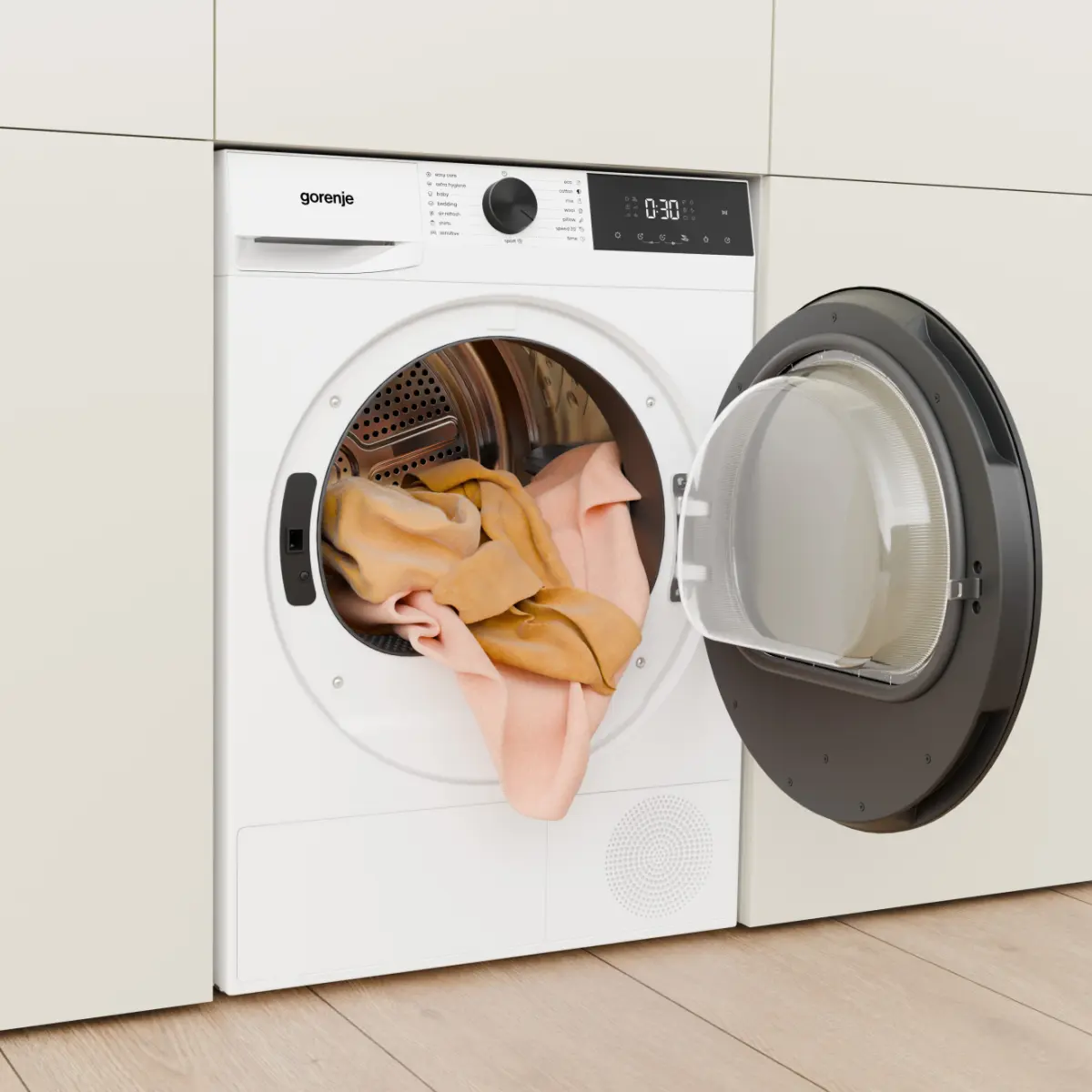 Egy fehér Gorenje szárítógép nyitott ajtóval, benne rózsaszín és sárga ruhákkal, világos fapadlón.