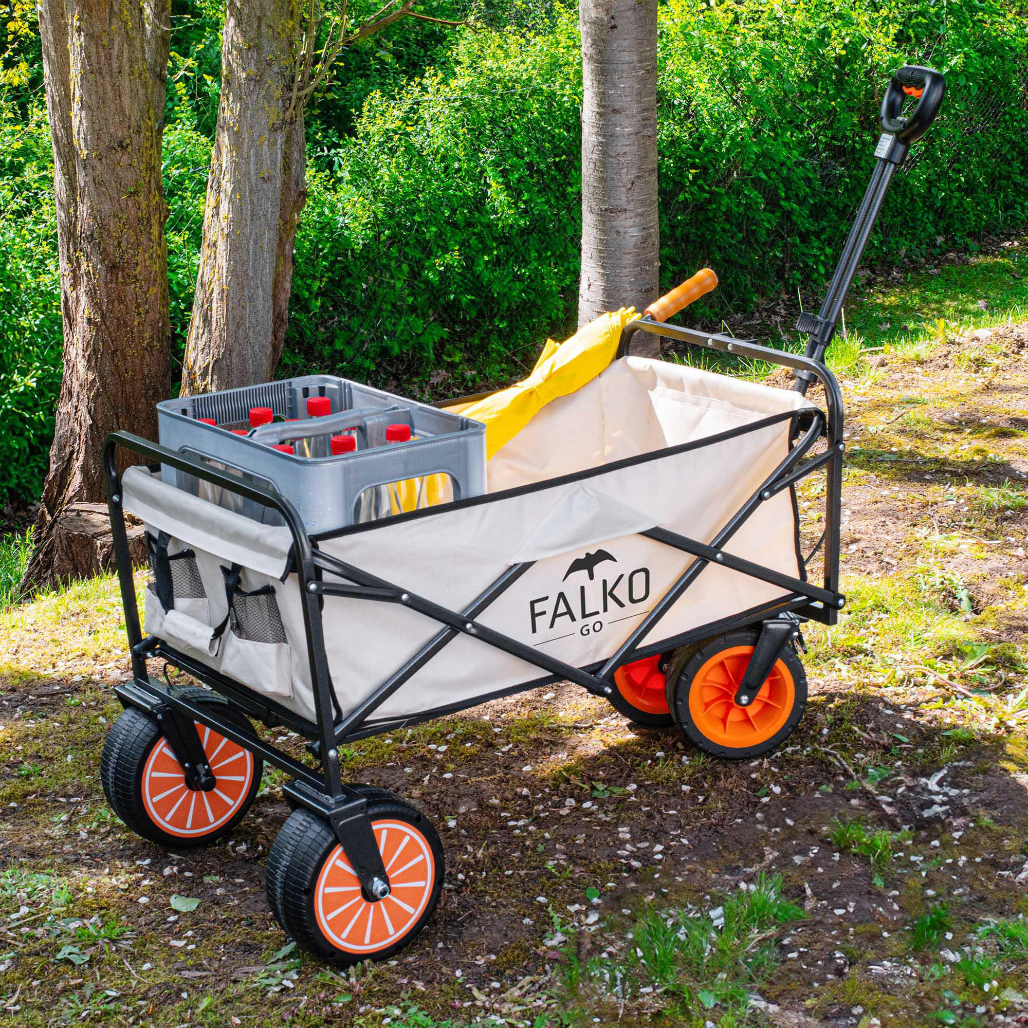 Ein beige Handwagen mit orangen Rädern und einem Getränkekasten steht im Wald auf grasigem Boden.