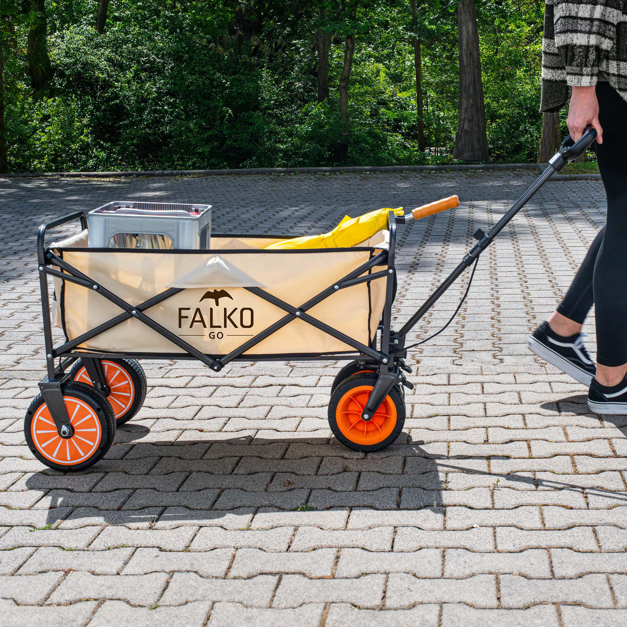 Eine Person zieht einen beige-schwarzen Handwagen mit Orangen-Rädern und Kiste auf Steinpflaster.