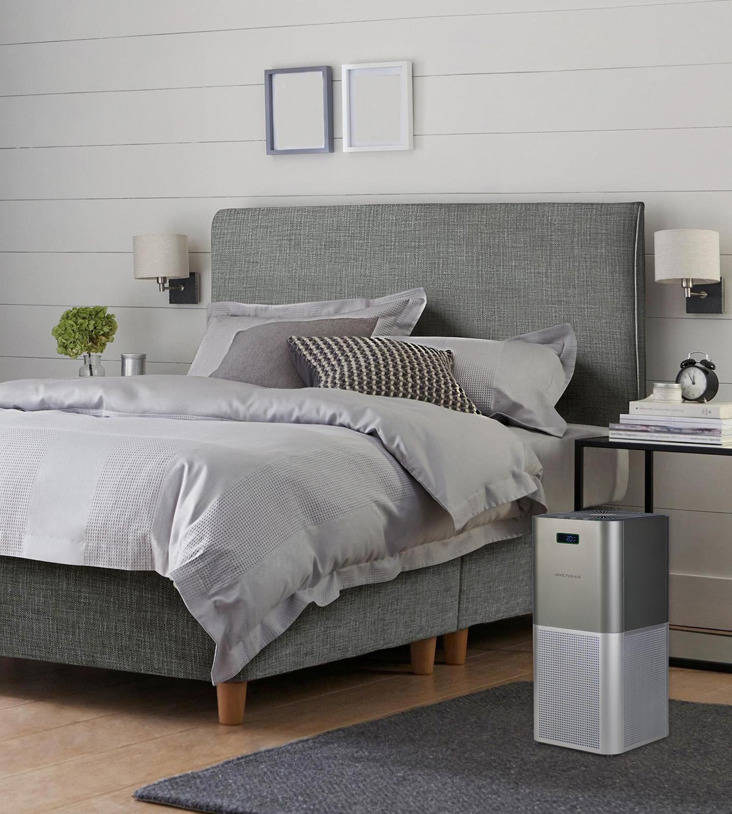 Ein grauer Luftreiniger steht im Schlafzimmer neben einem großen, grauen Bett vor einer weißen holzverkleideten Wand.
