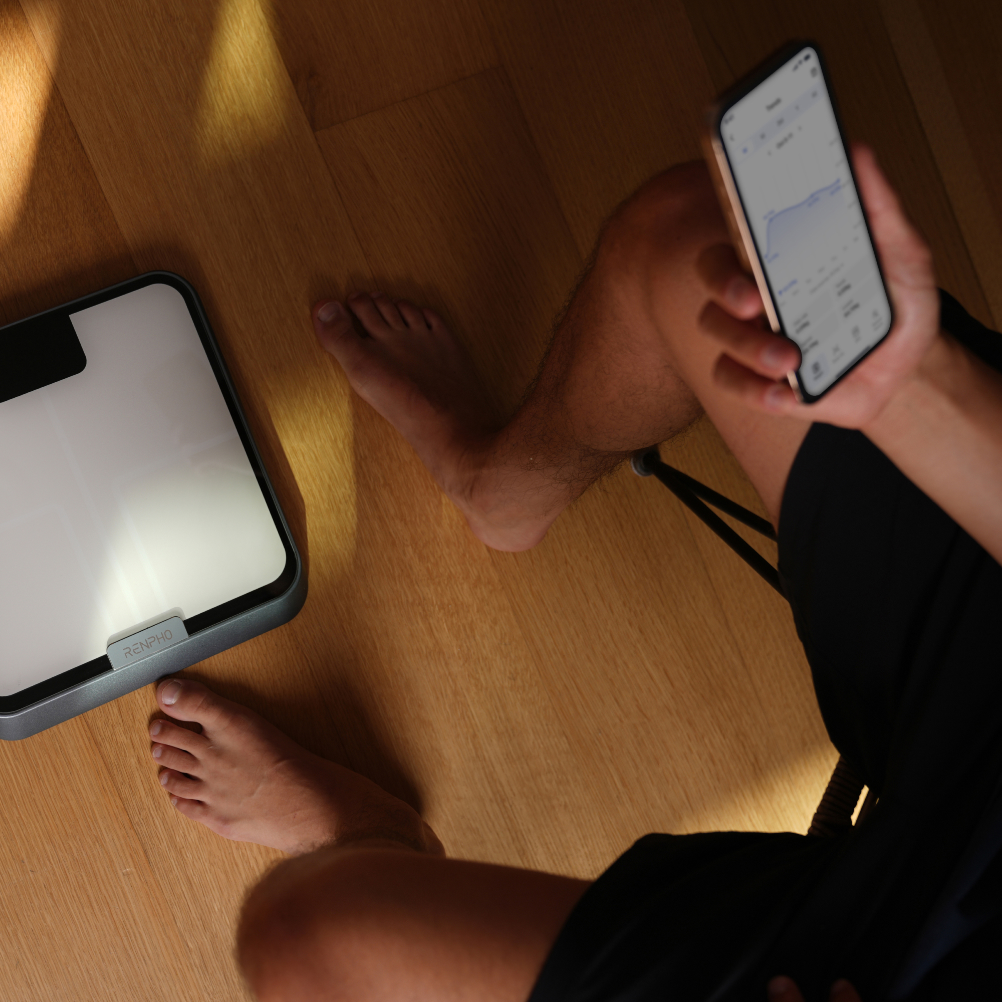 Persona mira descalza un teléfono inteligente con datos de salud junto a una báscula Renpho en suelo de madera.