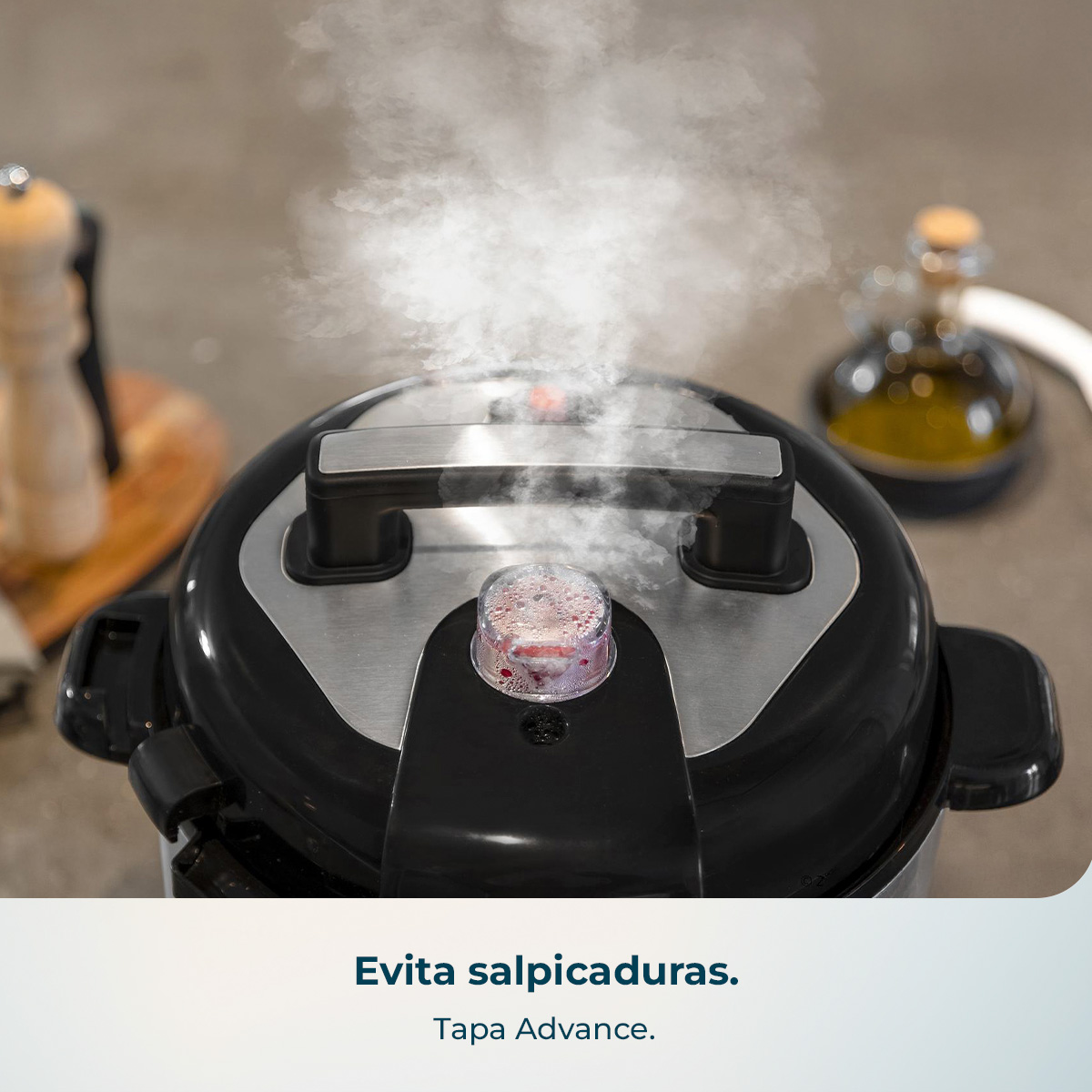 Una olla a presión negra emite vapor blanco espeso desde su válvula, con aceite y un molinillo al fondo.