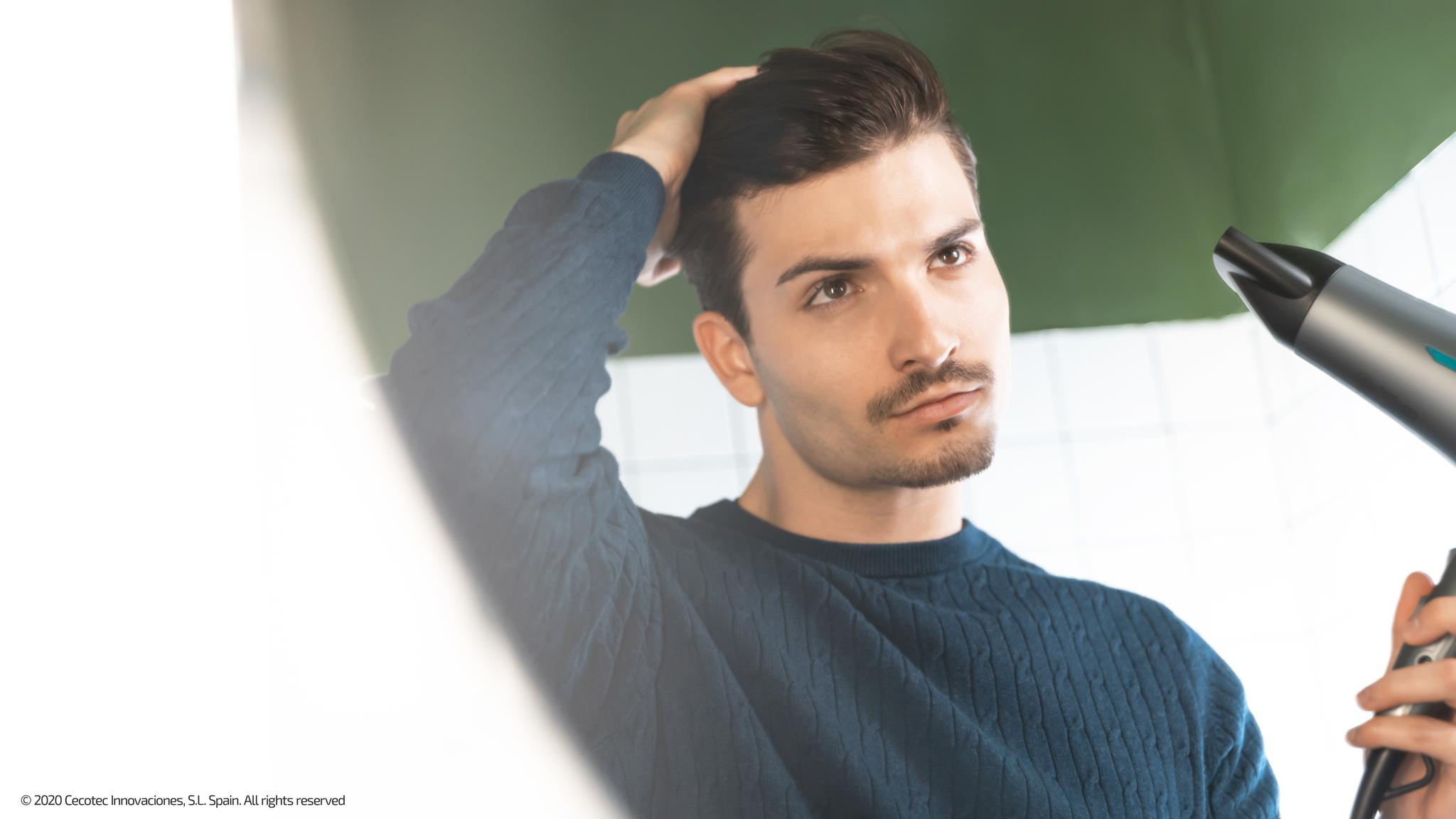 Un joven con jersey azul se peina el pelo oscuro con un secador frente a un espejo.