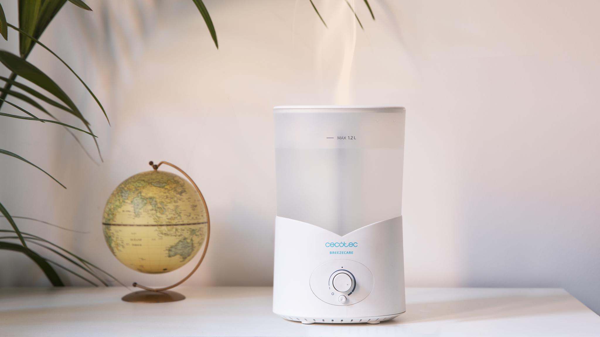 Un humidificador blanco con vapor ascendente sobre una mesa, con un globo terráqueo y hojas verdes al fondo.