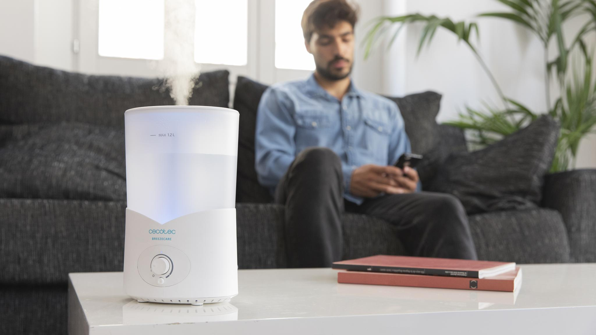Un humidificador blanco sobre una mesa, con un hombre sentado en el sofá al fondo.