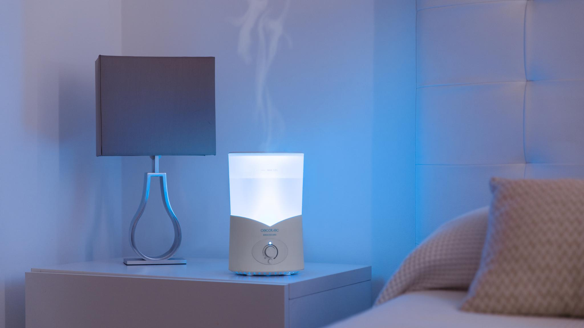 Un humidificador blanco con luz azul y vapor ascendente está en una mesita de noche junto a una lámpara.
