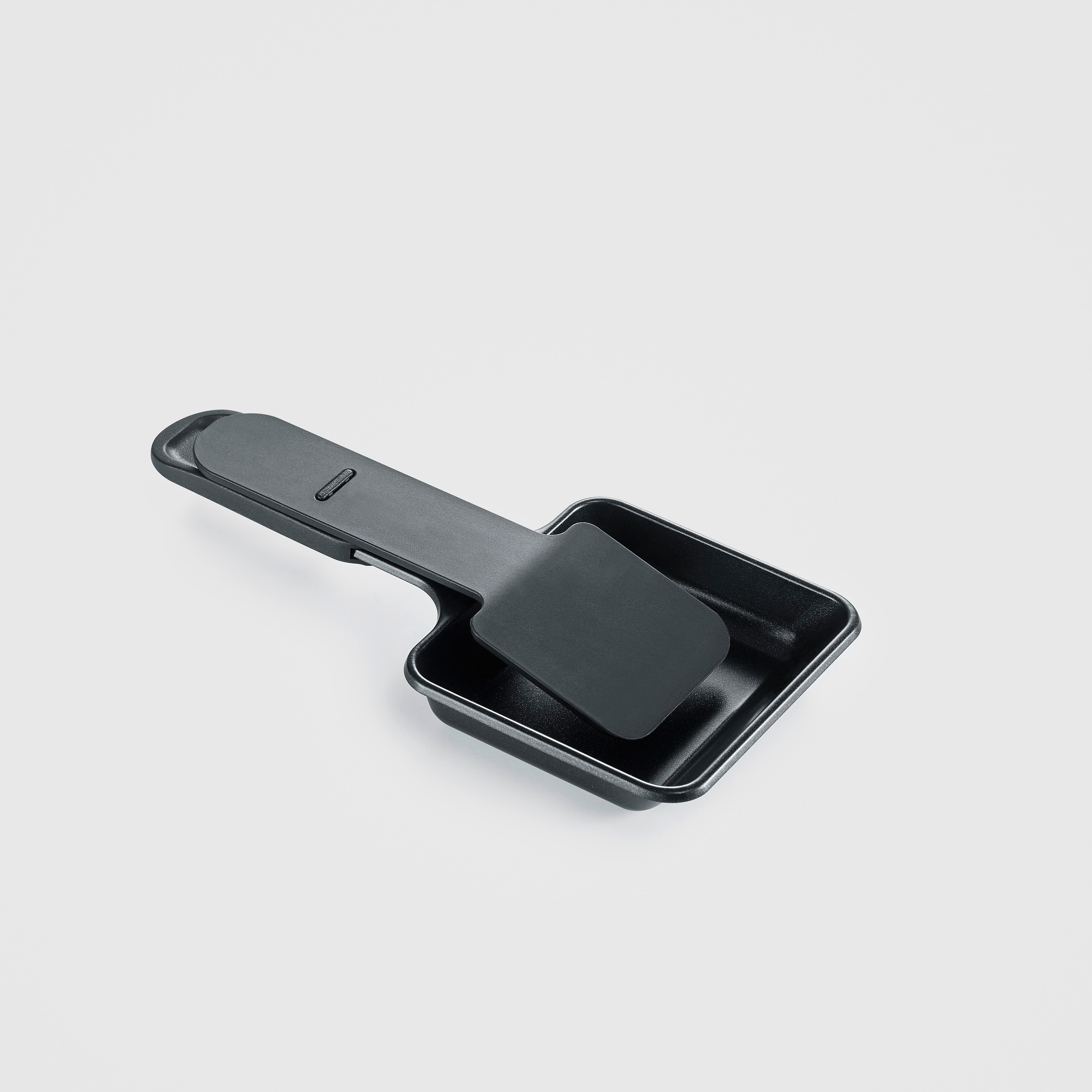Eine schwarze Raclette-Pfanne mit einem passenden Spatel darauf vor weißem Hintergrund.