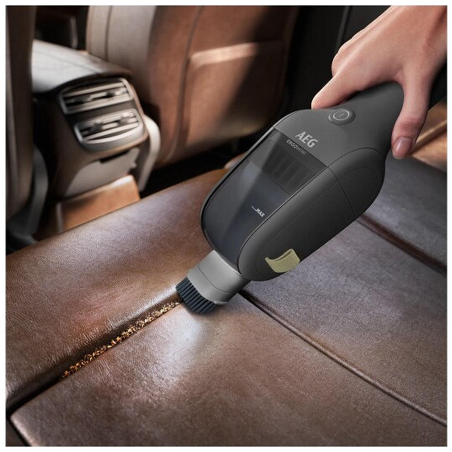 Een hand gebruikt een grijze AEG kruimeldief om een leren autostoelnaad te reinigen.
