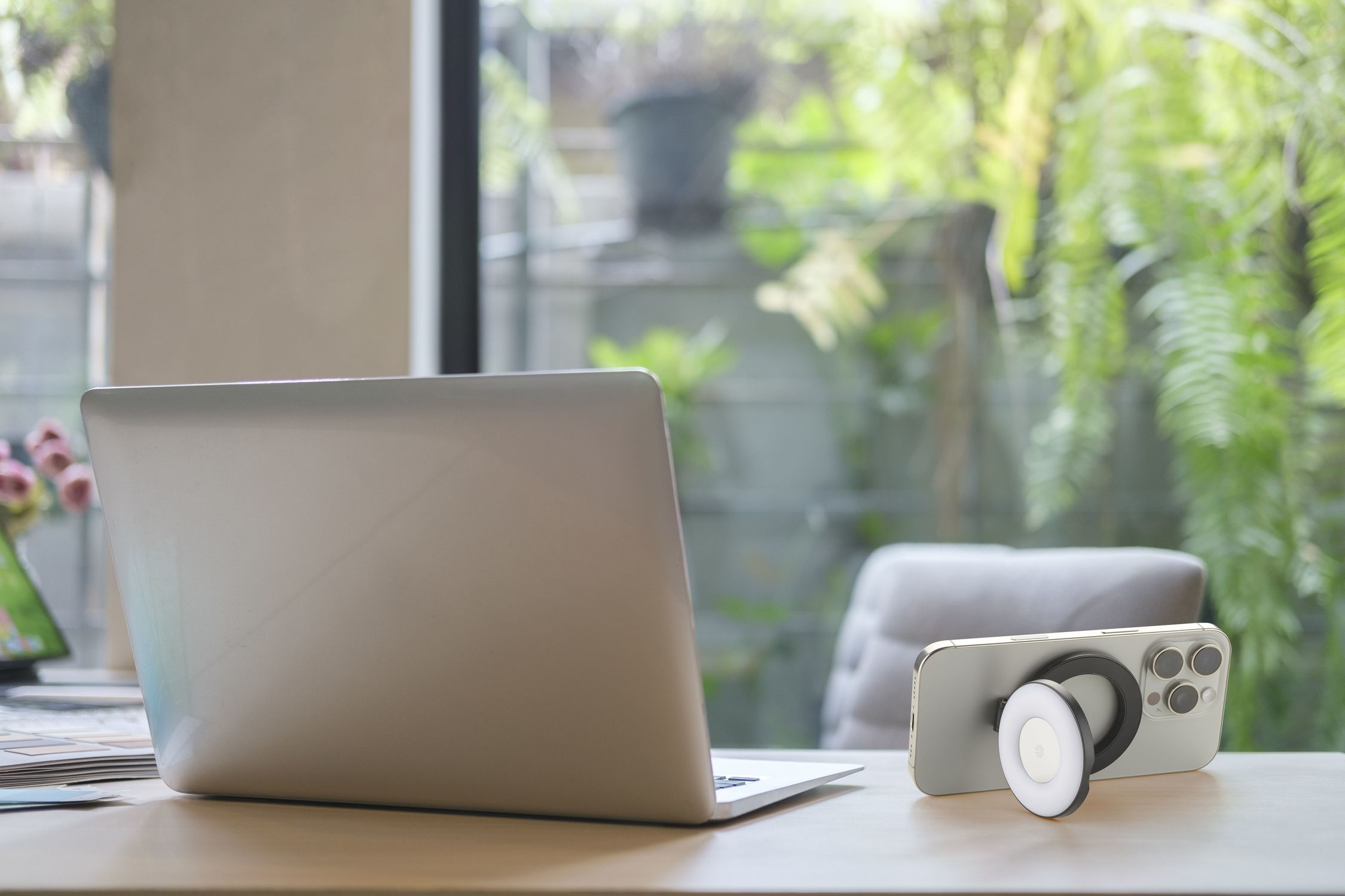 Una laptop y un smartphone con luz de aro magnética sobre un escritorio de madera con vista al jardín.