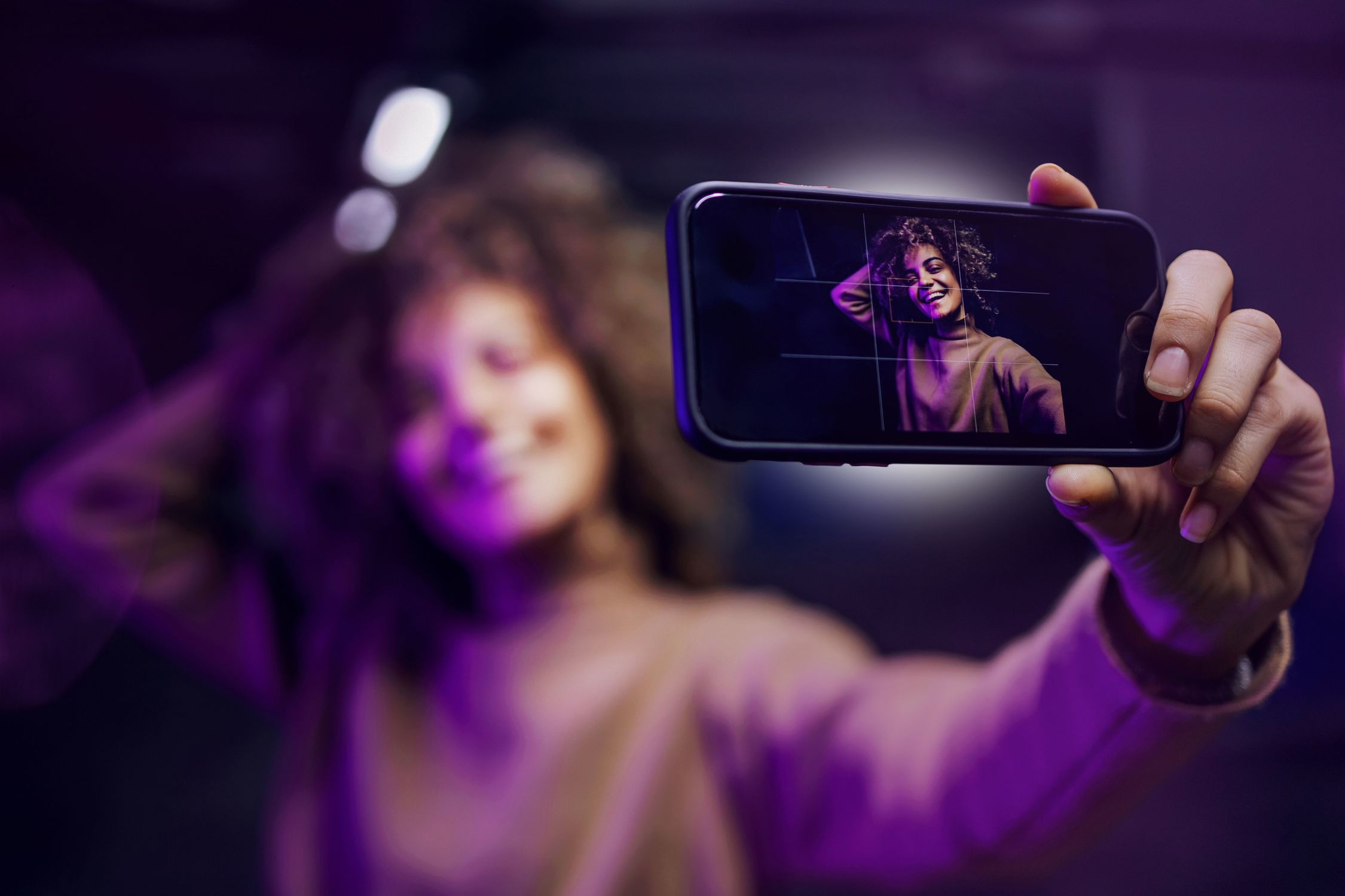 Una mujer con cabello rizado sostiene un teléfono inteligente y toma una selfie bajo iluminación púrpura.