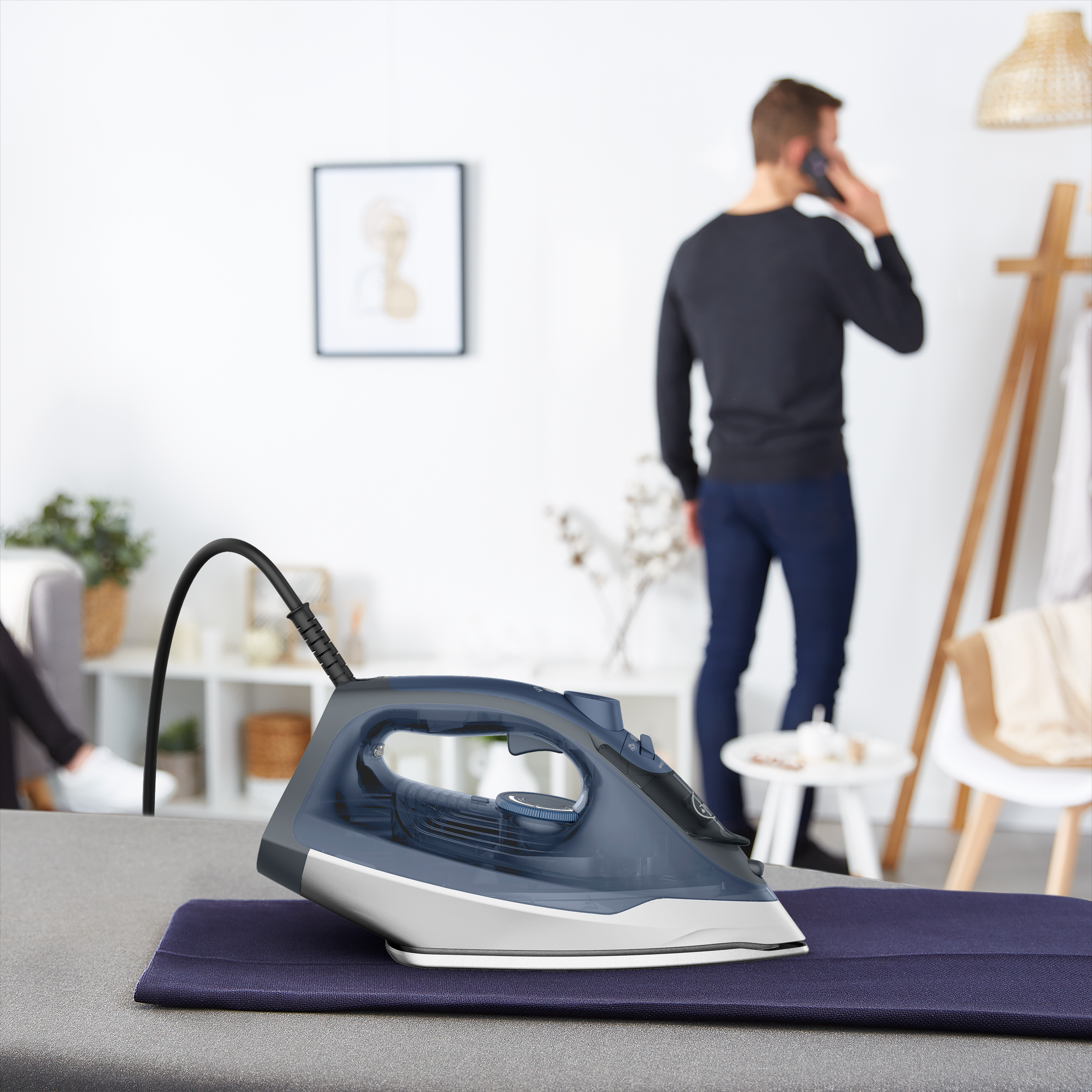 Egy modern gőzölős vasaló áll egy alátéten, miközben a háttérben egy férfi telefonál.
