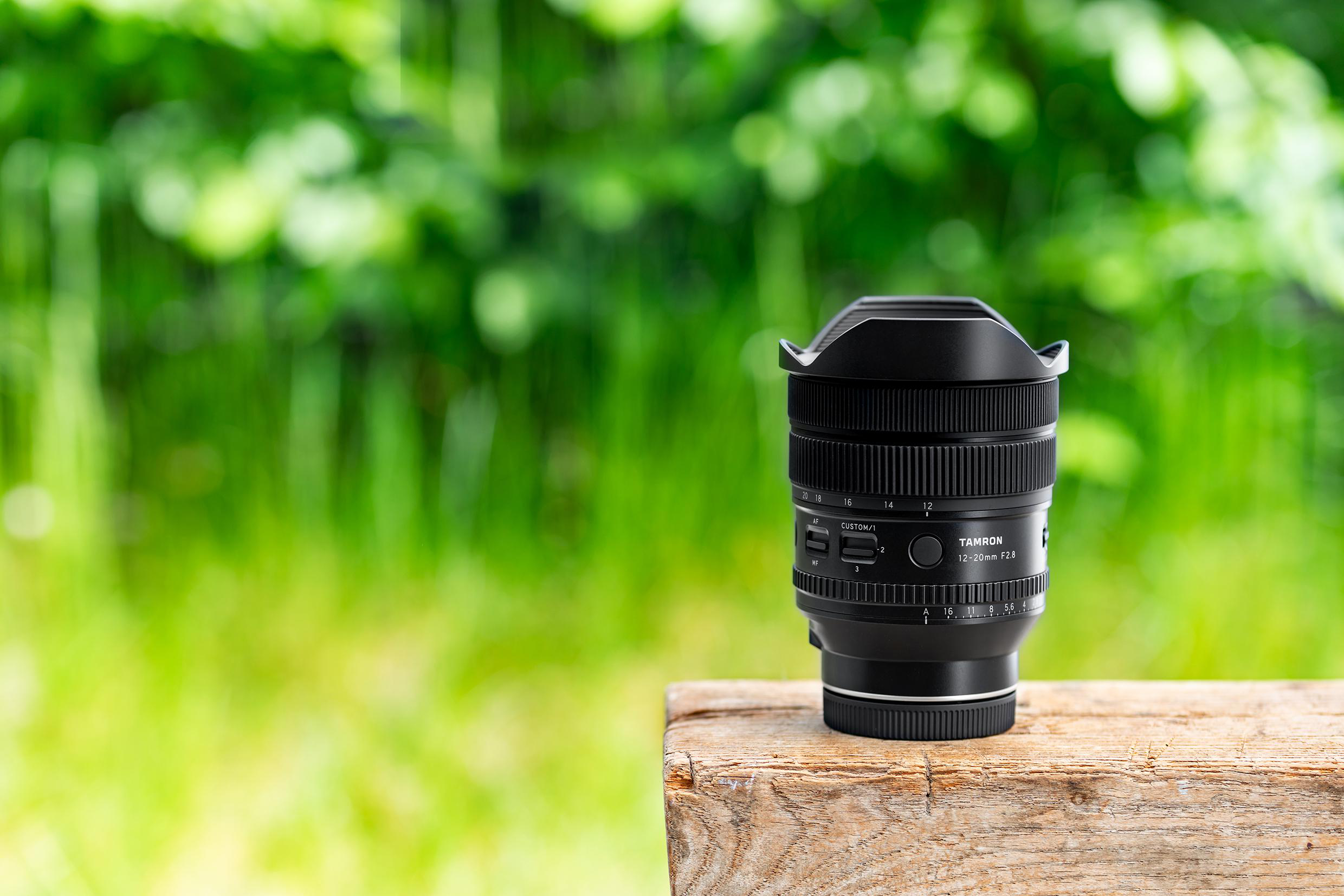 Ein schwarzes Tamron 12-20mm F2.8 Kameraobjektiv steht auf einem Holzblock vor grünem Hintergrund.