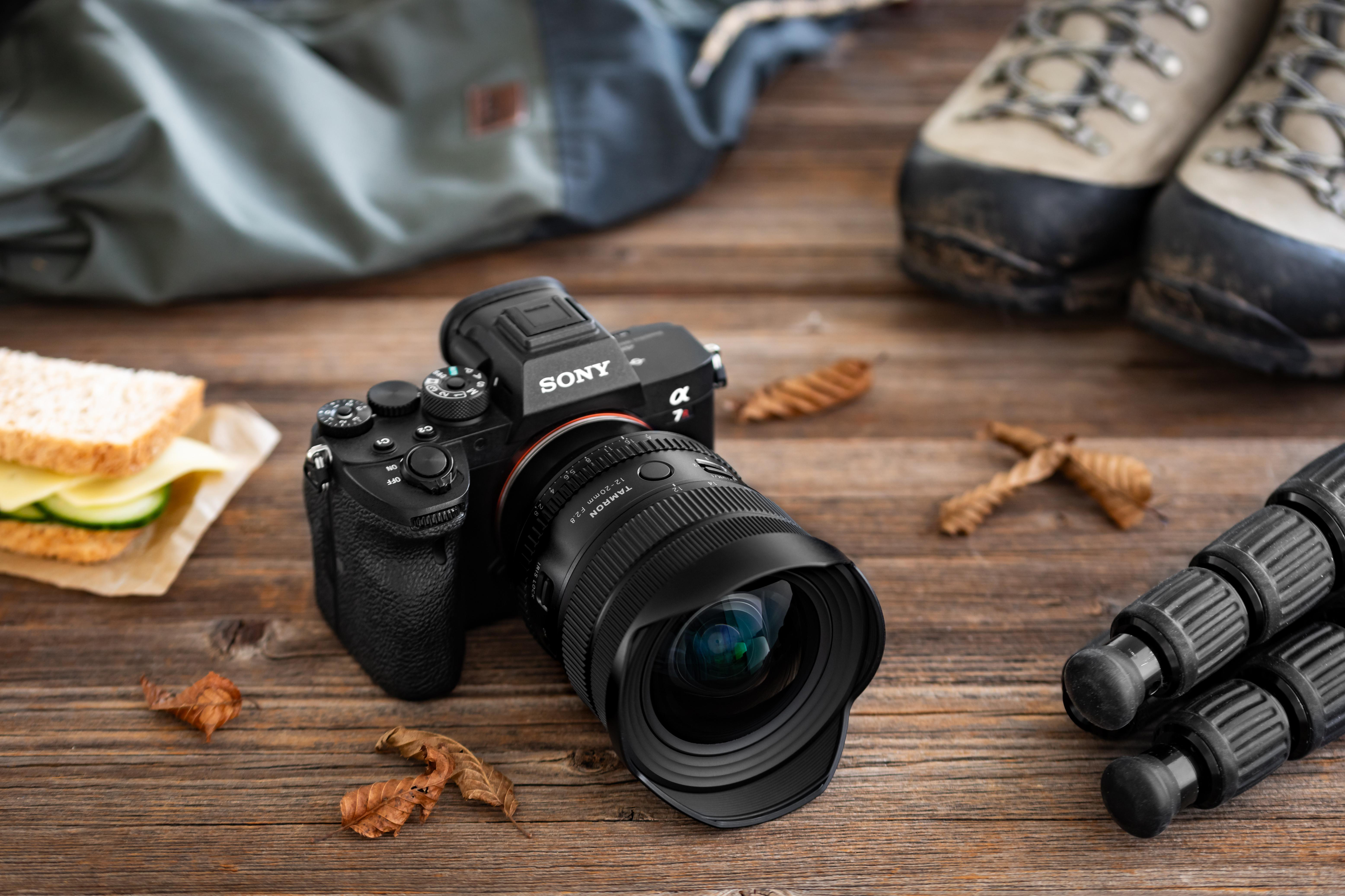 Eine Sony-Kamera mit Tamron-Objektiv, Wanderschuhe, Rucksack und ein Sandwich liegen auf einem Holztisch.