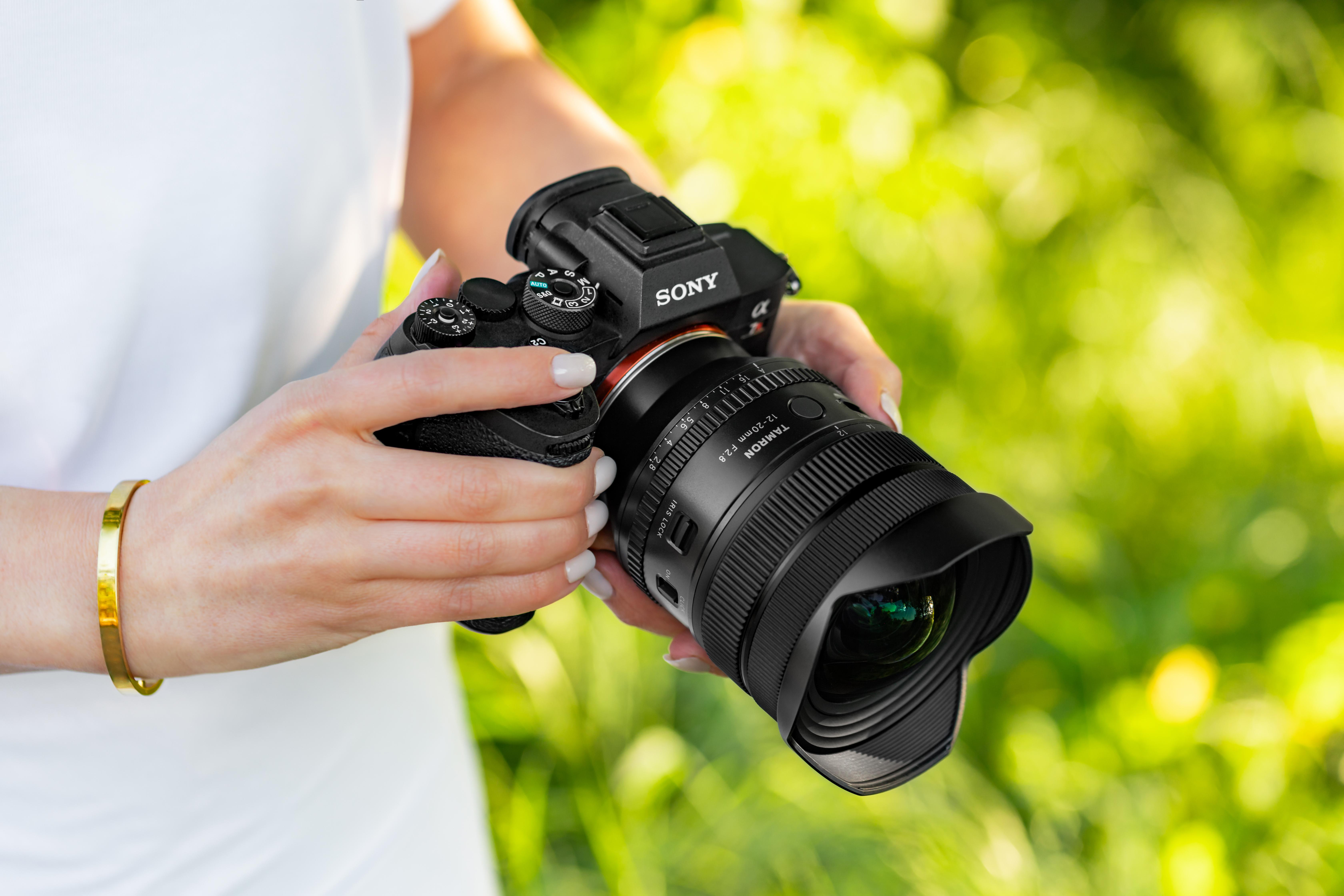 Eine Frau hält eine Sony-Kamera mit einem Tamron-Objektiv vor grünem Hintergrund.