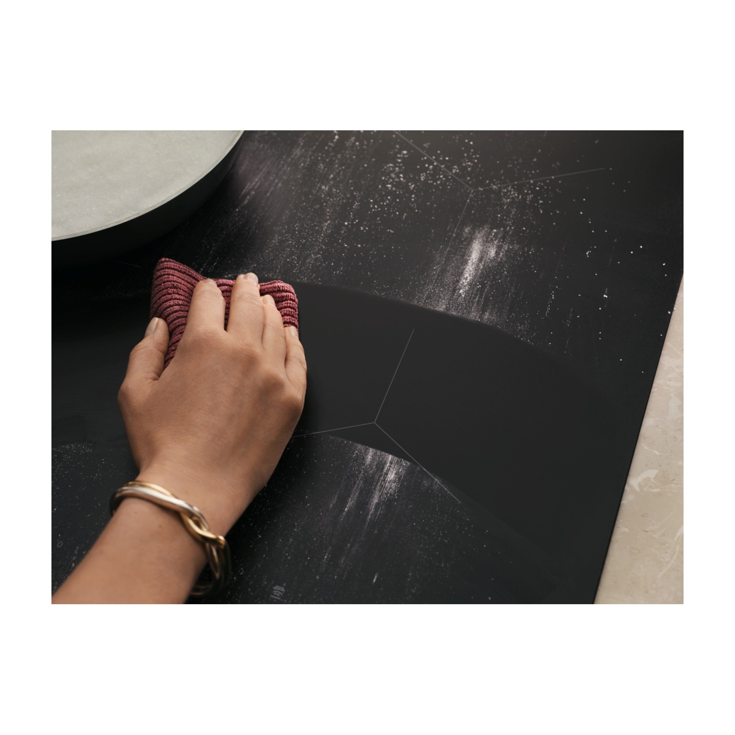 Una mano con un braccialetto pulisce un piano cottura a induzione nero spolverato di farina con un panno rosso.