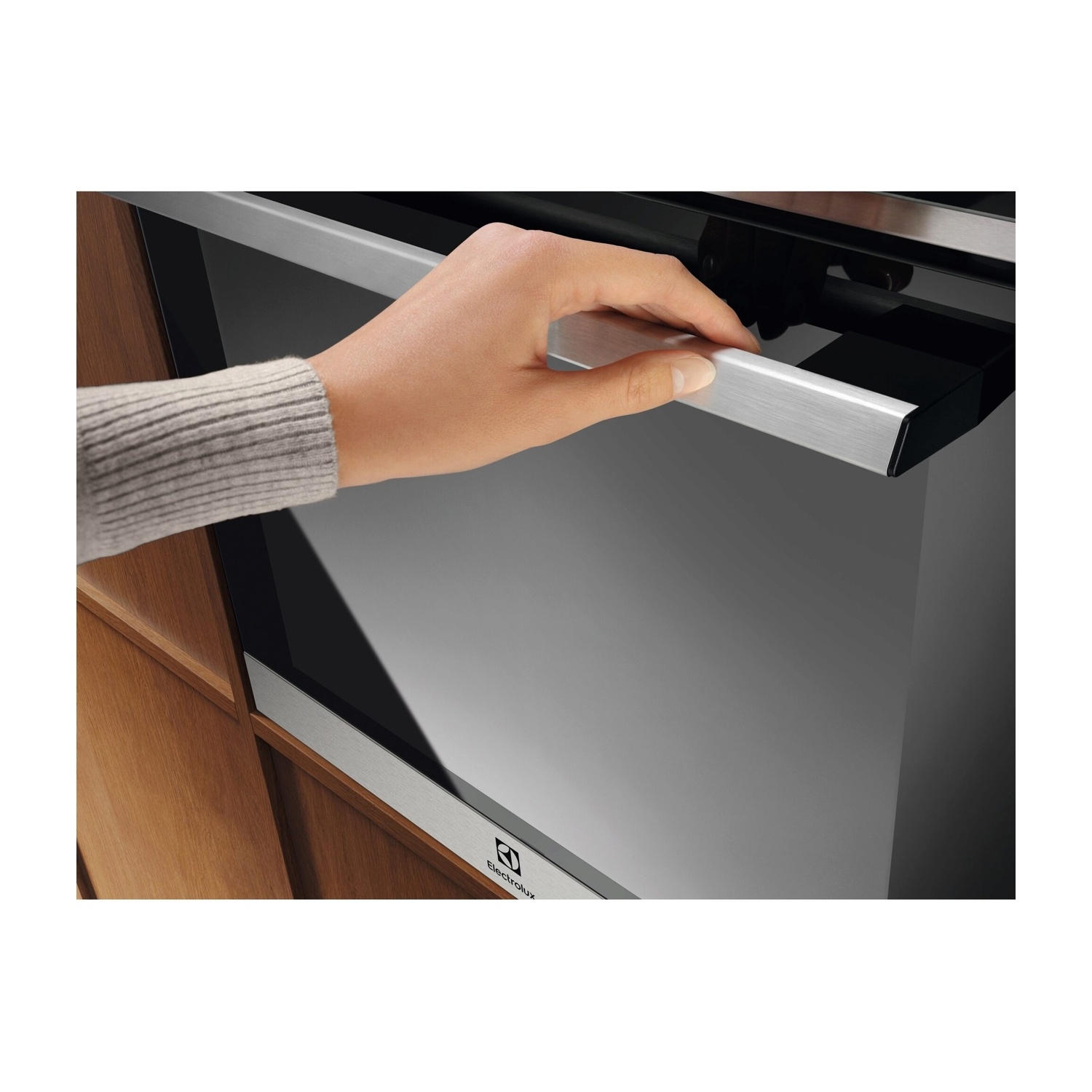 Una mano apre la maniglia argentata di un moderno forno Electrolux nero incassato in mobili in legno.