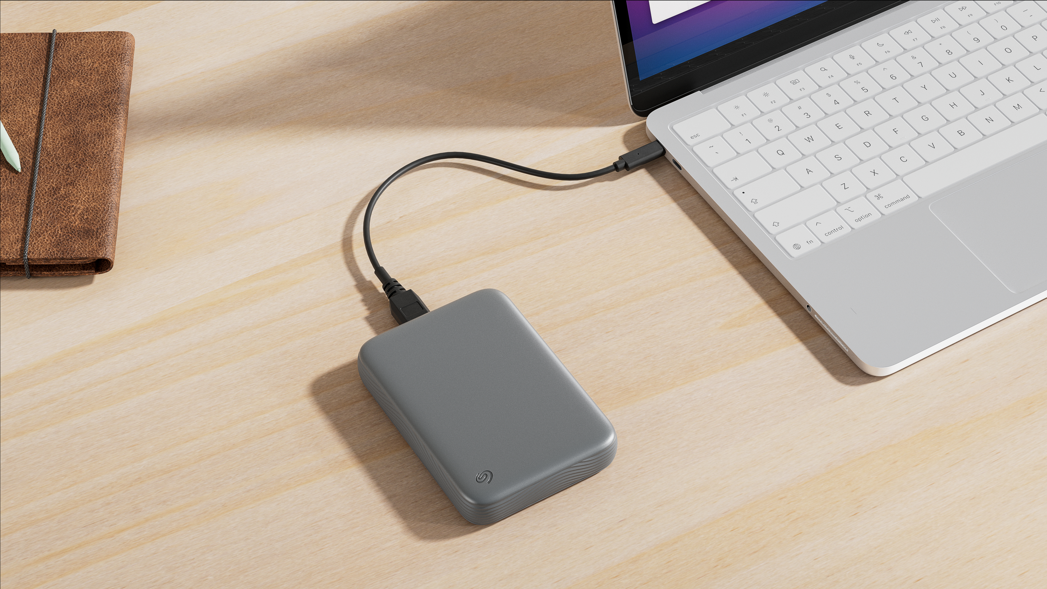 Eine graue externe Festplatte, verbunden mit einem Laptop auf einem Holztisch mit einem braunen Notizbuch.