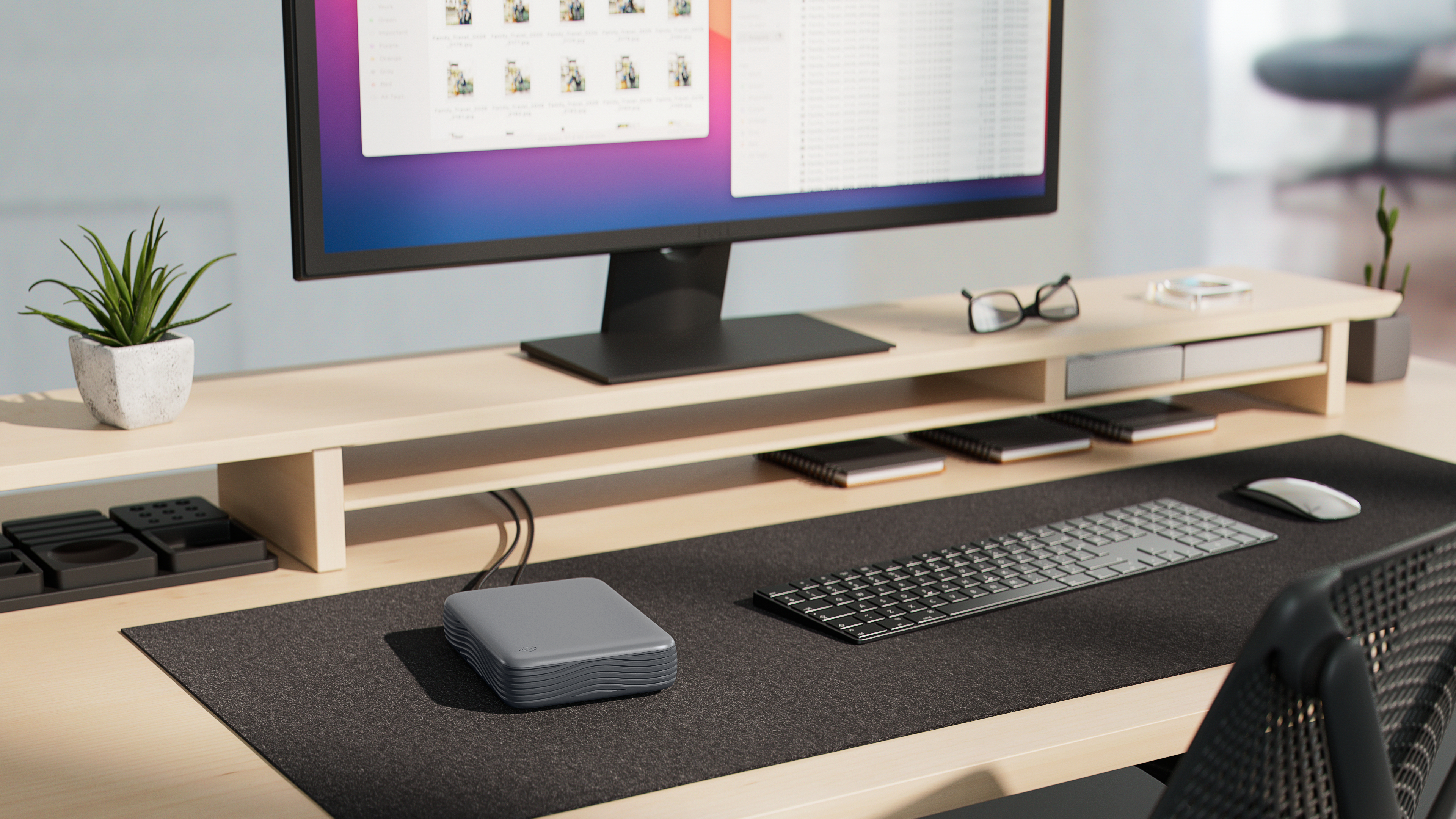 Ein moderner Holztisch mit Monitor, Tastatur, Maus, Pflanzen und grauer externer Festplatte auf einer schwarzen Matte.