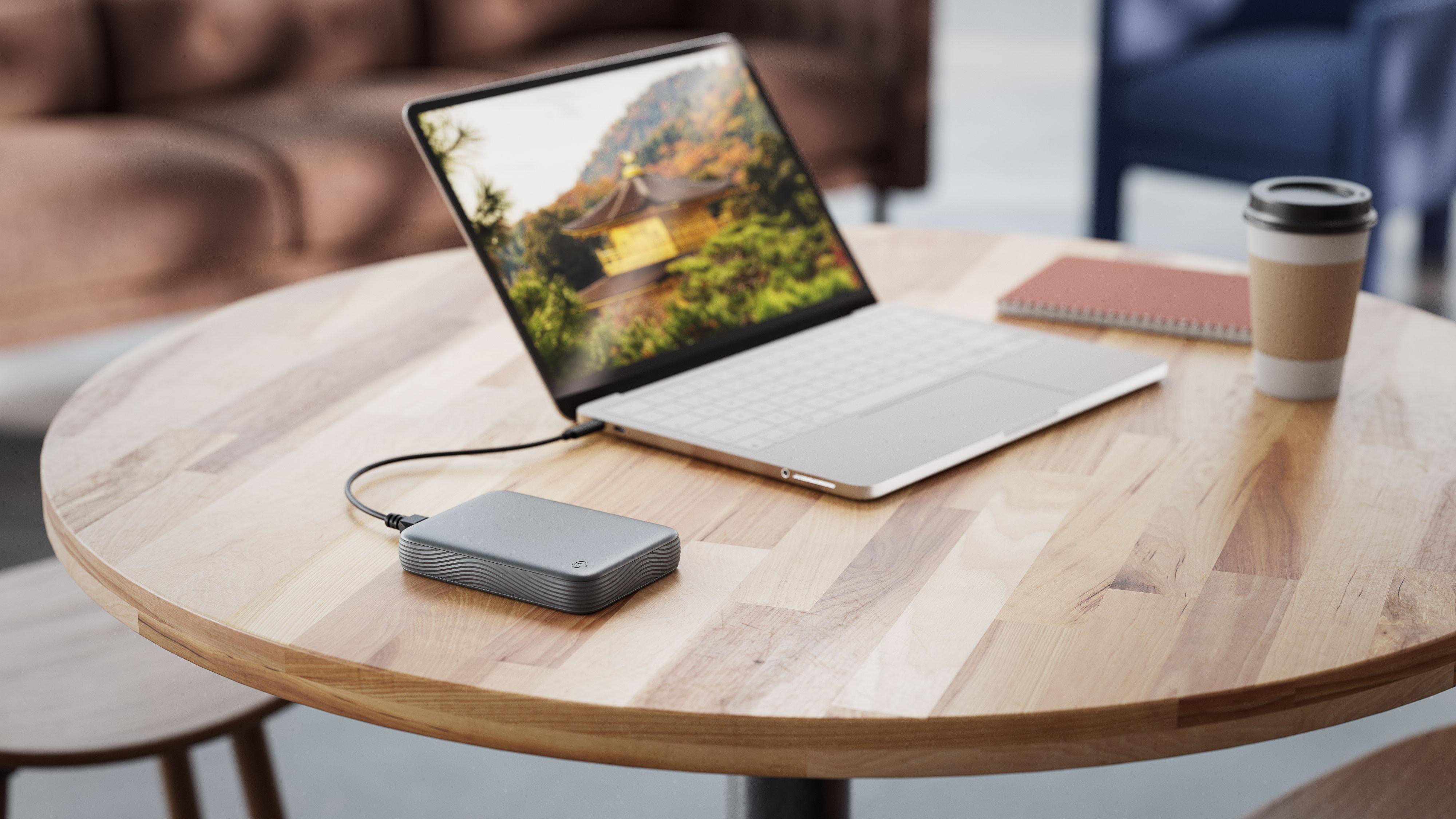 Eine graue externe Festplatte, verbunden mit einem Laptop auf einem Holztisch, neben Kaffee und Notizbuch.