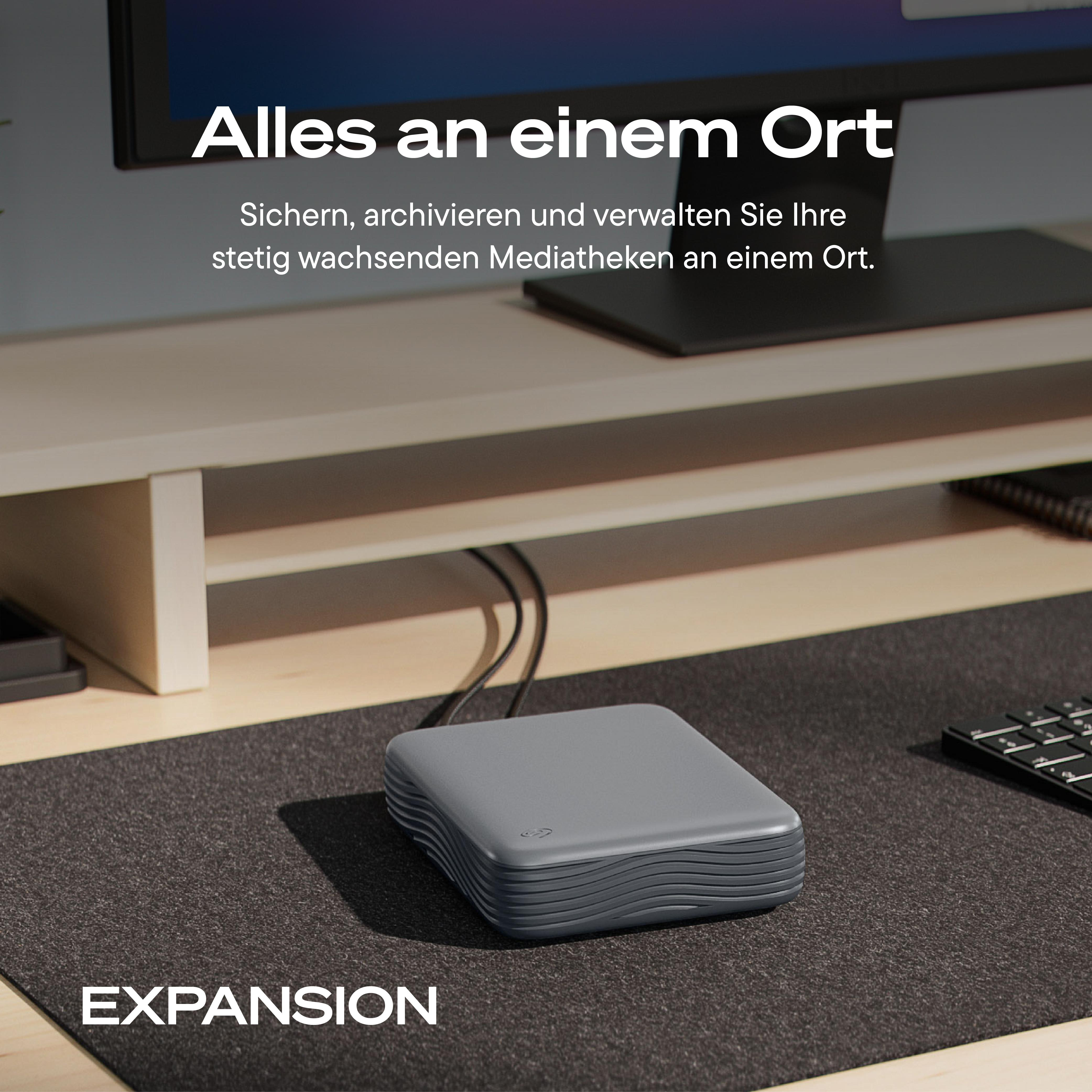 Eine graue Seagate-Festplatte steht auf einem Schreibtisch vor einem Monitor mit deutschem Werbetext.