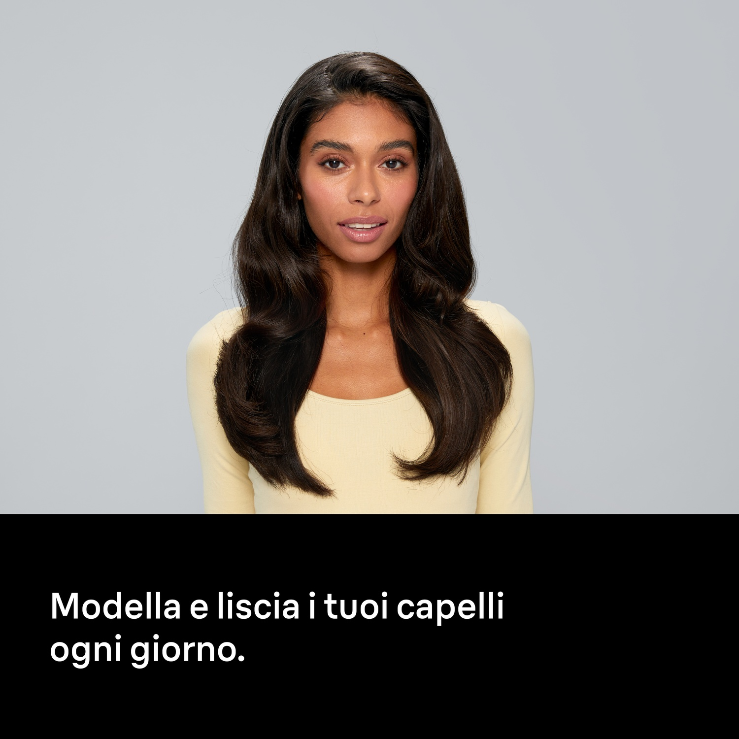 Una donna con capelli lunghi, scuri e mossi e un top giallo su sfondo grigio chiaro.