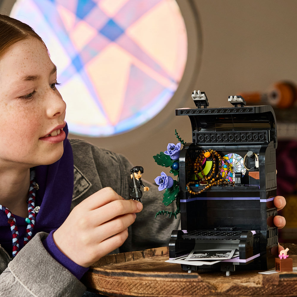 Ein Mädchen hält eine LEGO Mittwoch-Figur vor ein offenes, schwarz-lila Modell mit blauem Blumenmuster.