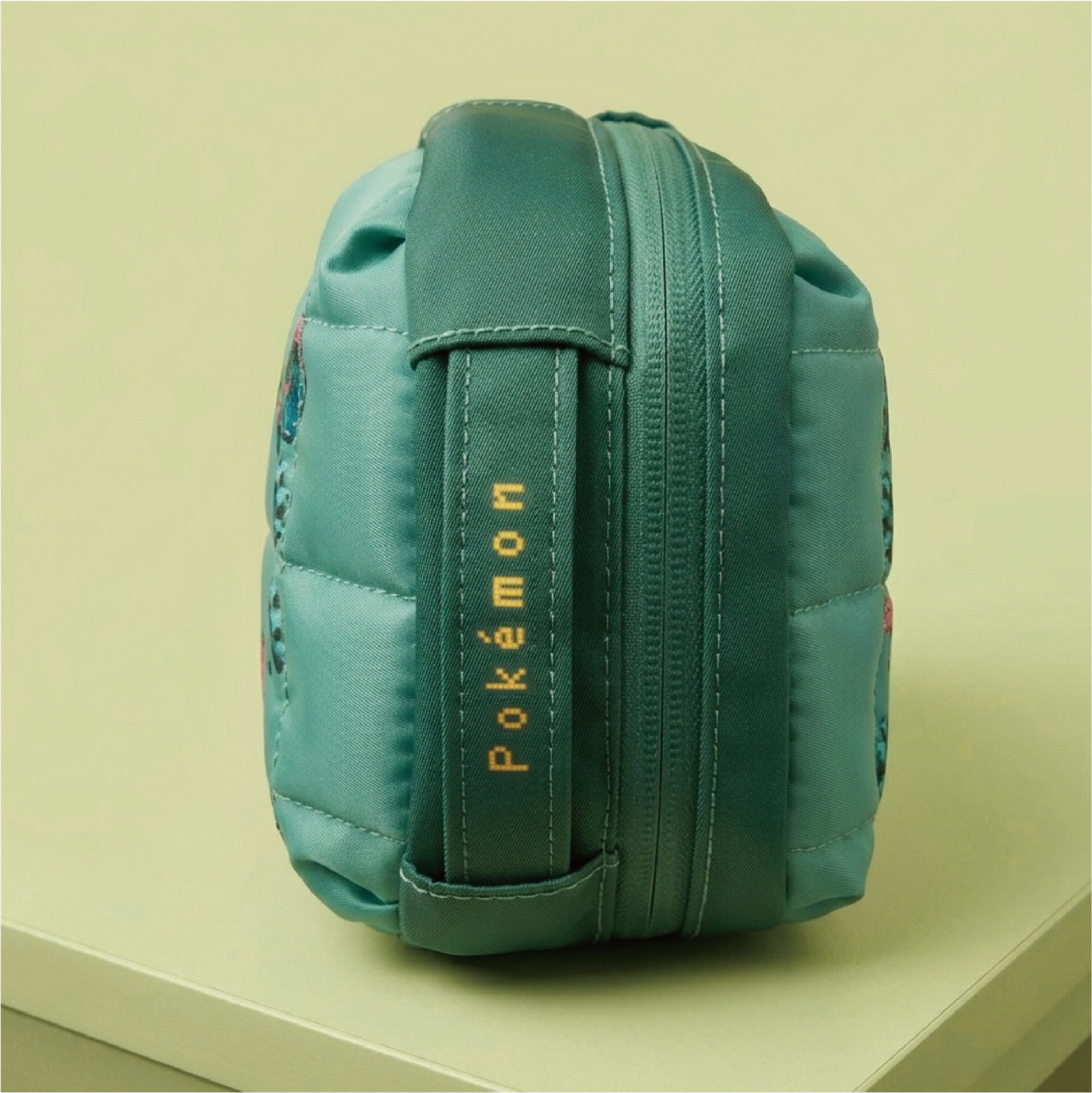 Una borsa verde con cerniera Pokémon è appoggiata su una superficie verde chiaro.