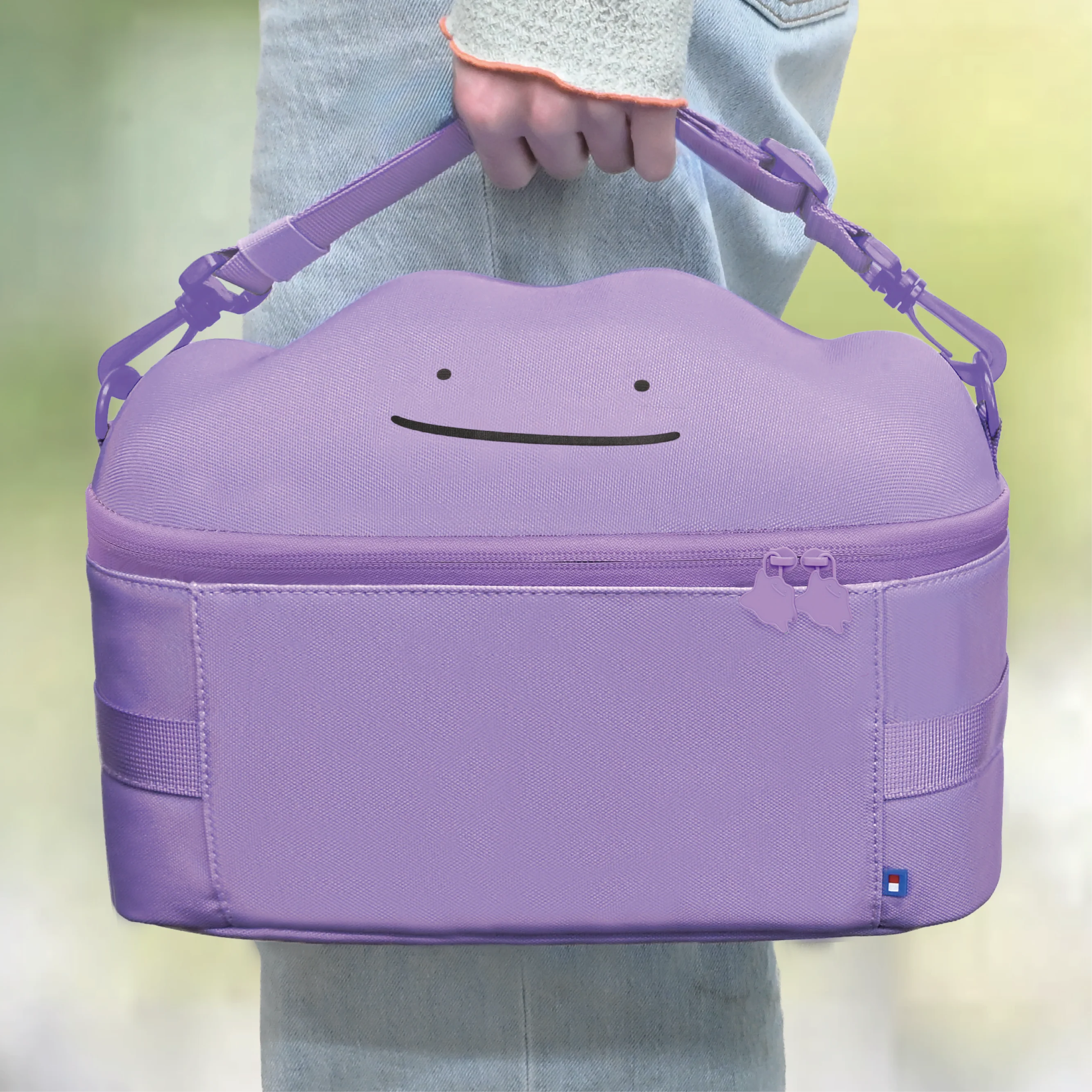 Una persona in jeans tiene in mano una borsa viola con il viso di Ditto stampato e cerniere.