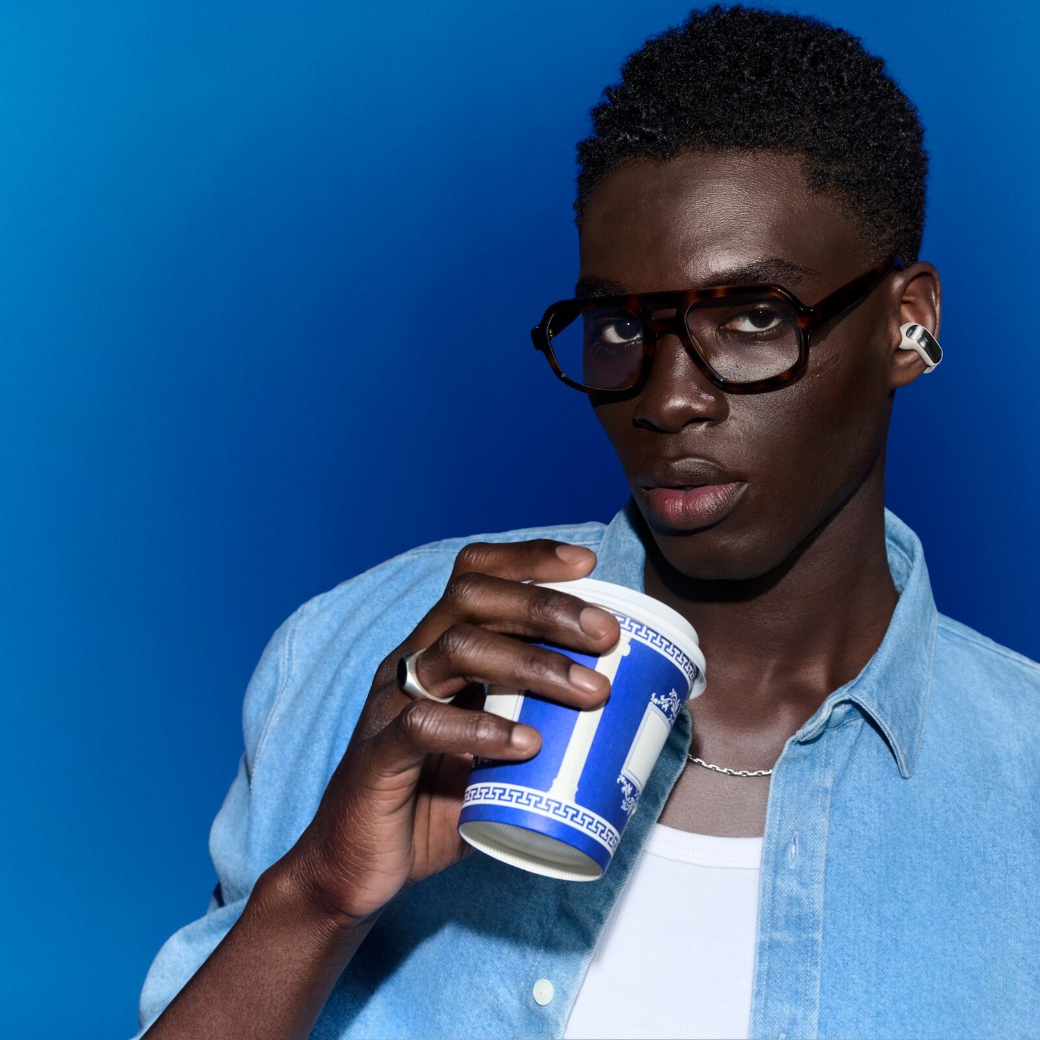 Een jonge man met een bril en een spijkerhemel drinkt uit een blauwe papieren beker tegen een blauwe achtergrond.
