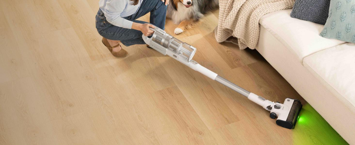 Eine Frau saugt mit einem kabellosen Staubsauger den Holzboden vor einem Sofa, während ein Hund zuschaut.