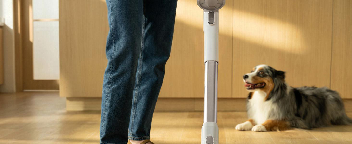 Eine Person mit Jeans nutzt einen Staubsauger, während im Hintergrund ein Hund auf dem Holzboden liegt.