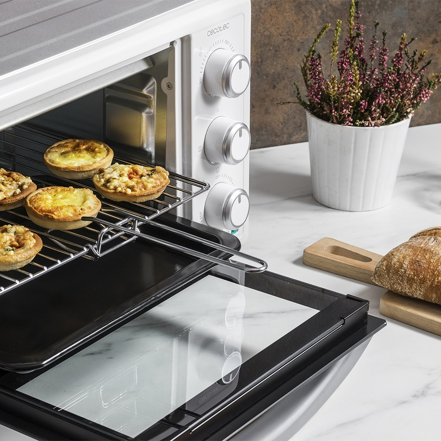 Un forno bianco con la porta aperta cuoce delle crostatine su un piano di lavoro accanto a pane e una pianta.