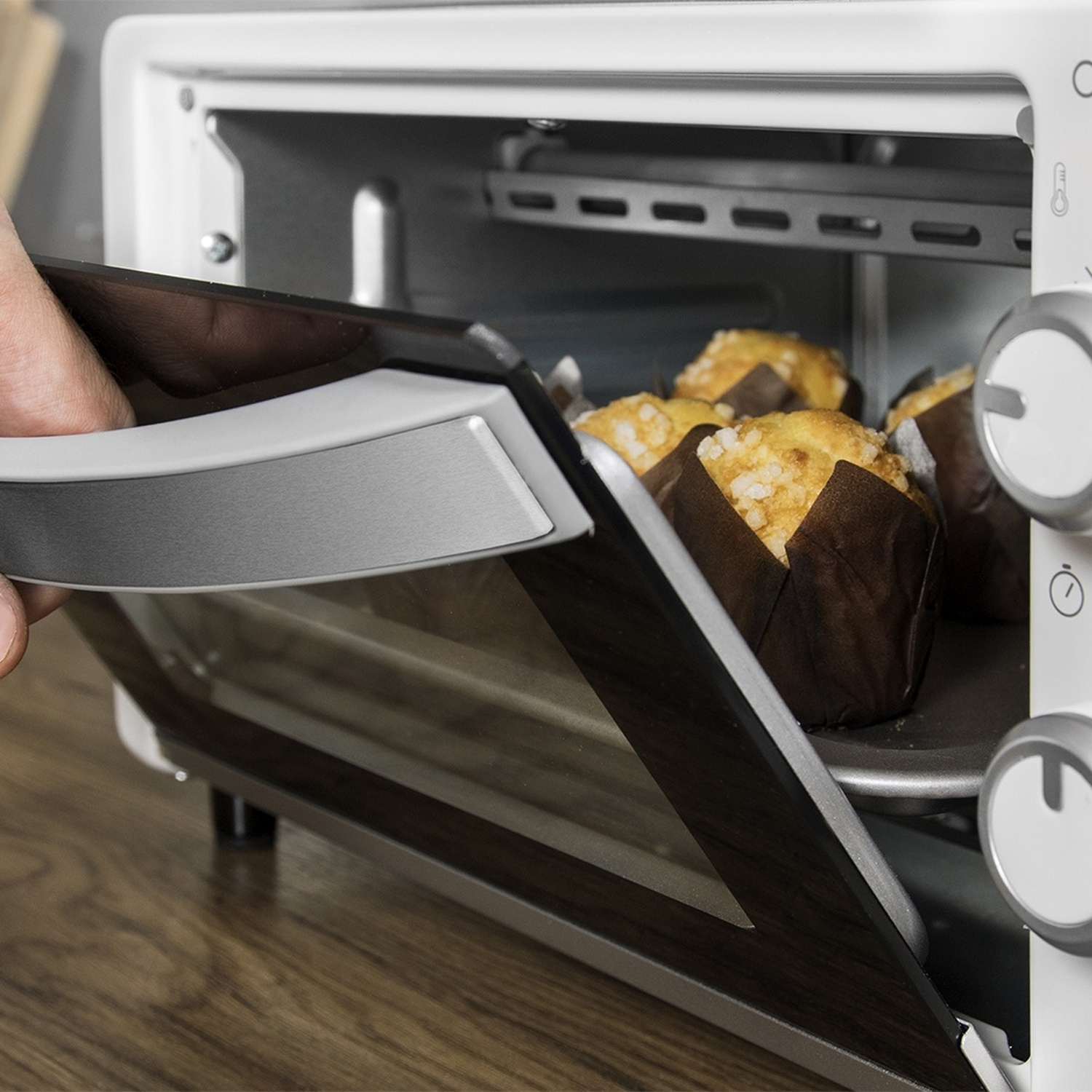 Una mano apre la porta a vetri di un mini forno bianco che cuoce muffin.