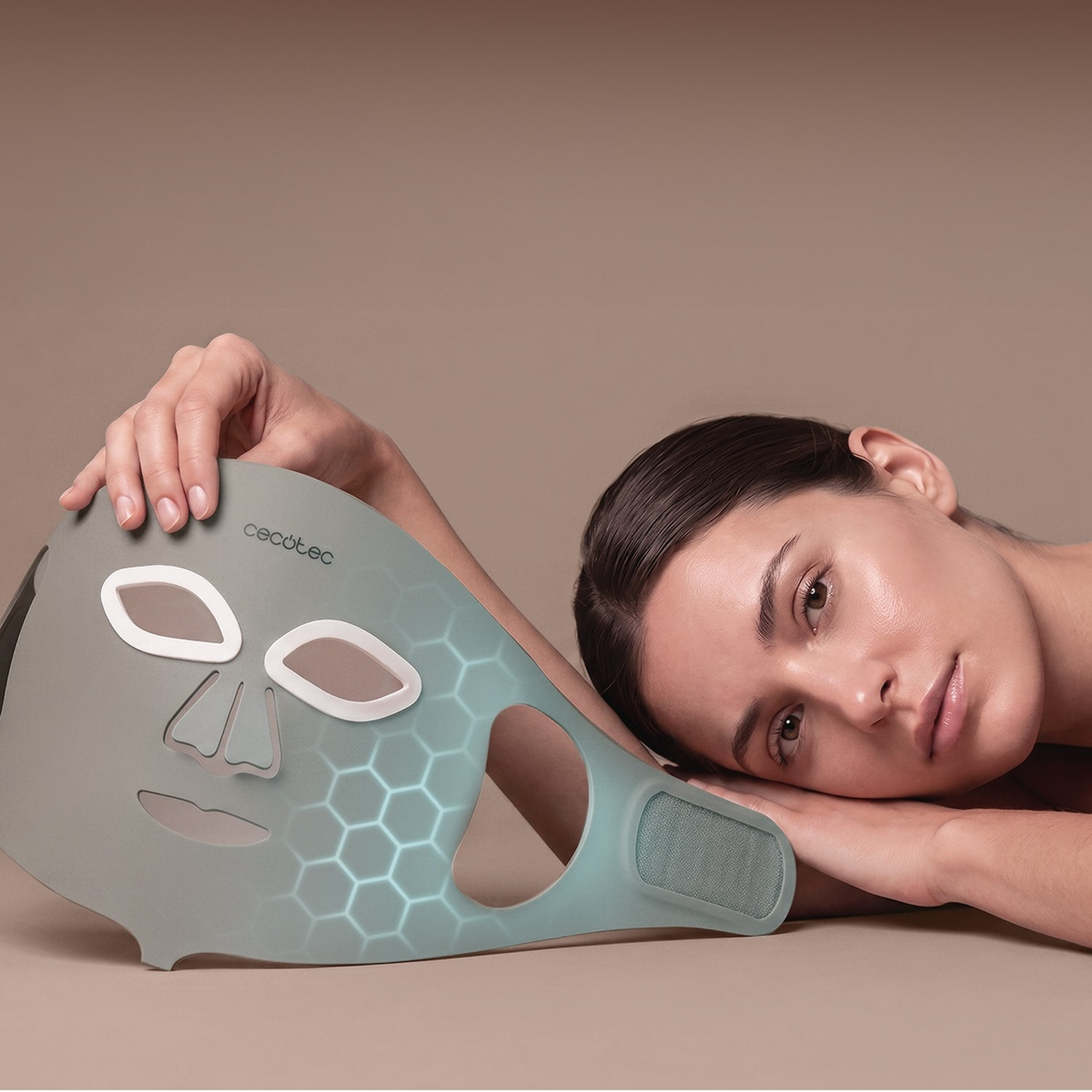 Una donna tiene in mano una maschera facciale LED grigia e luminosa su sfondo neutro.