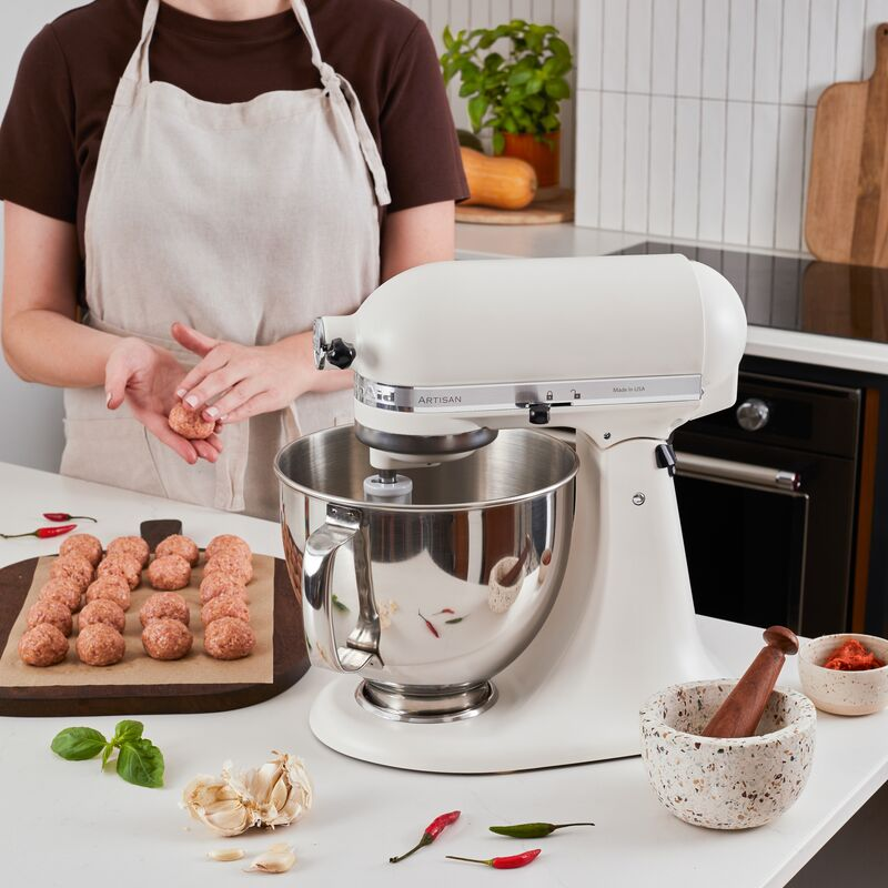 Person formt Fleischbällchen neben weißer Küchenmaschine, Gewürzen und Mörser.