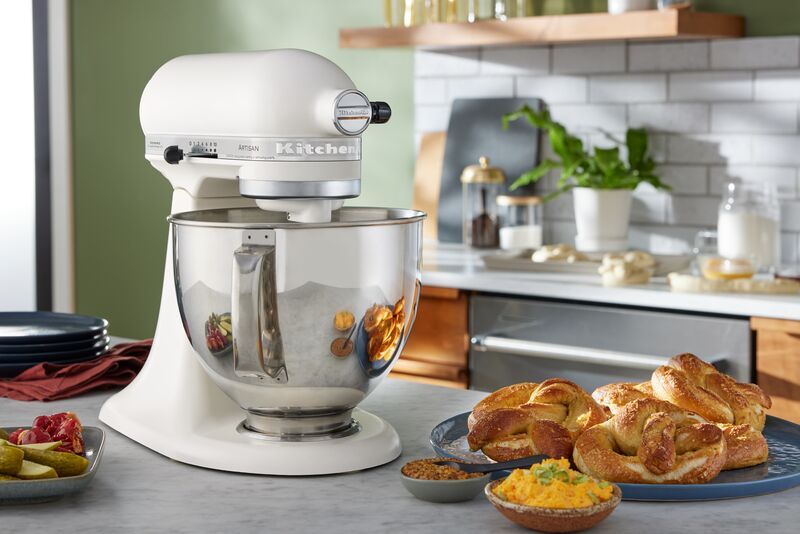 Eine weiße Küchenmaschine mit Edelstahlschüssel steht auf einer Theke neben frischen Brezeln und Dips.