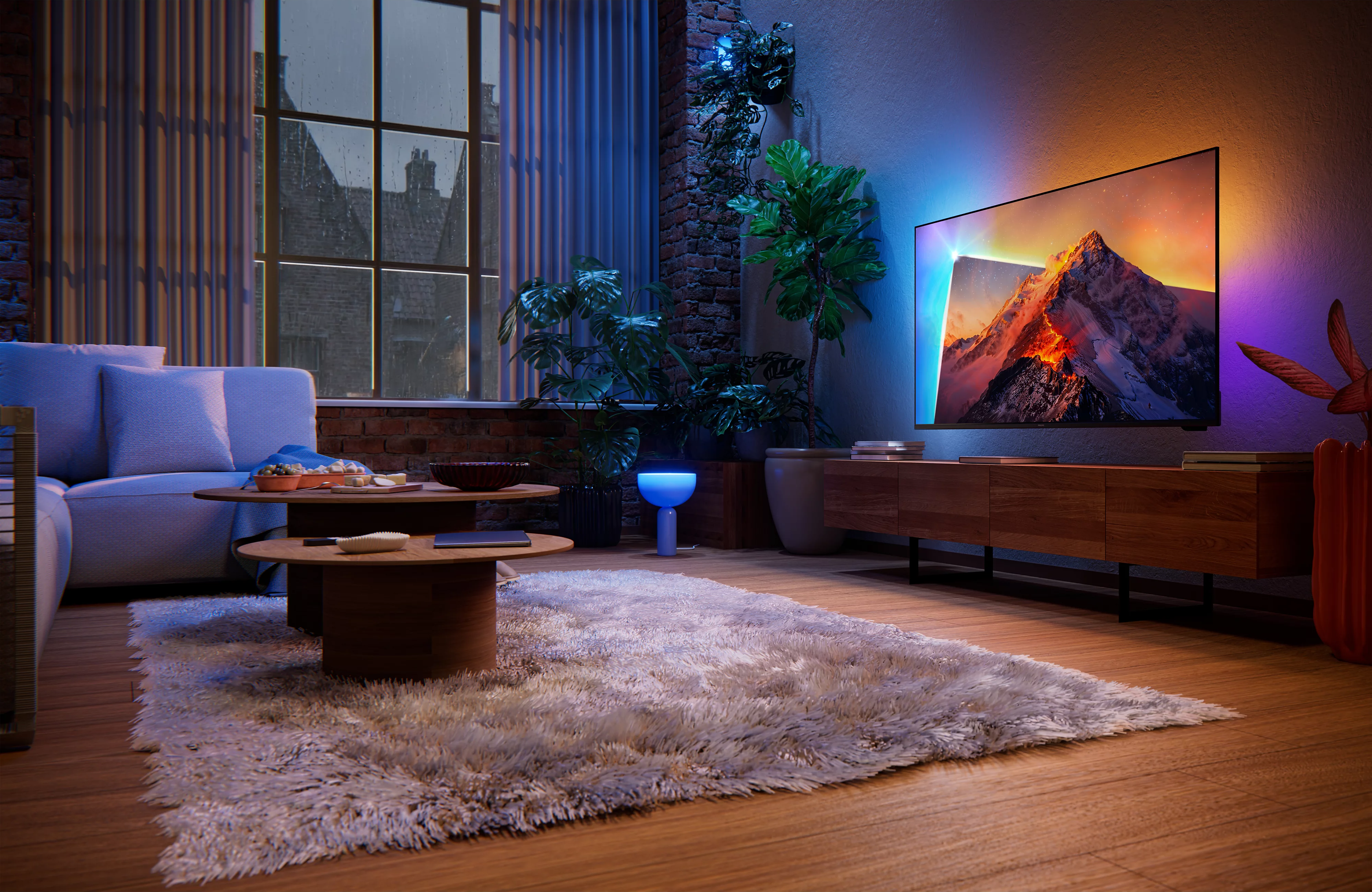 Salon de nuit avec canapé, table en bois, plantes et télévision sur le mur de briques.