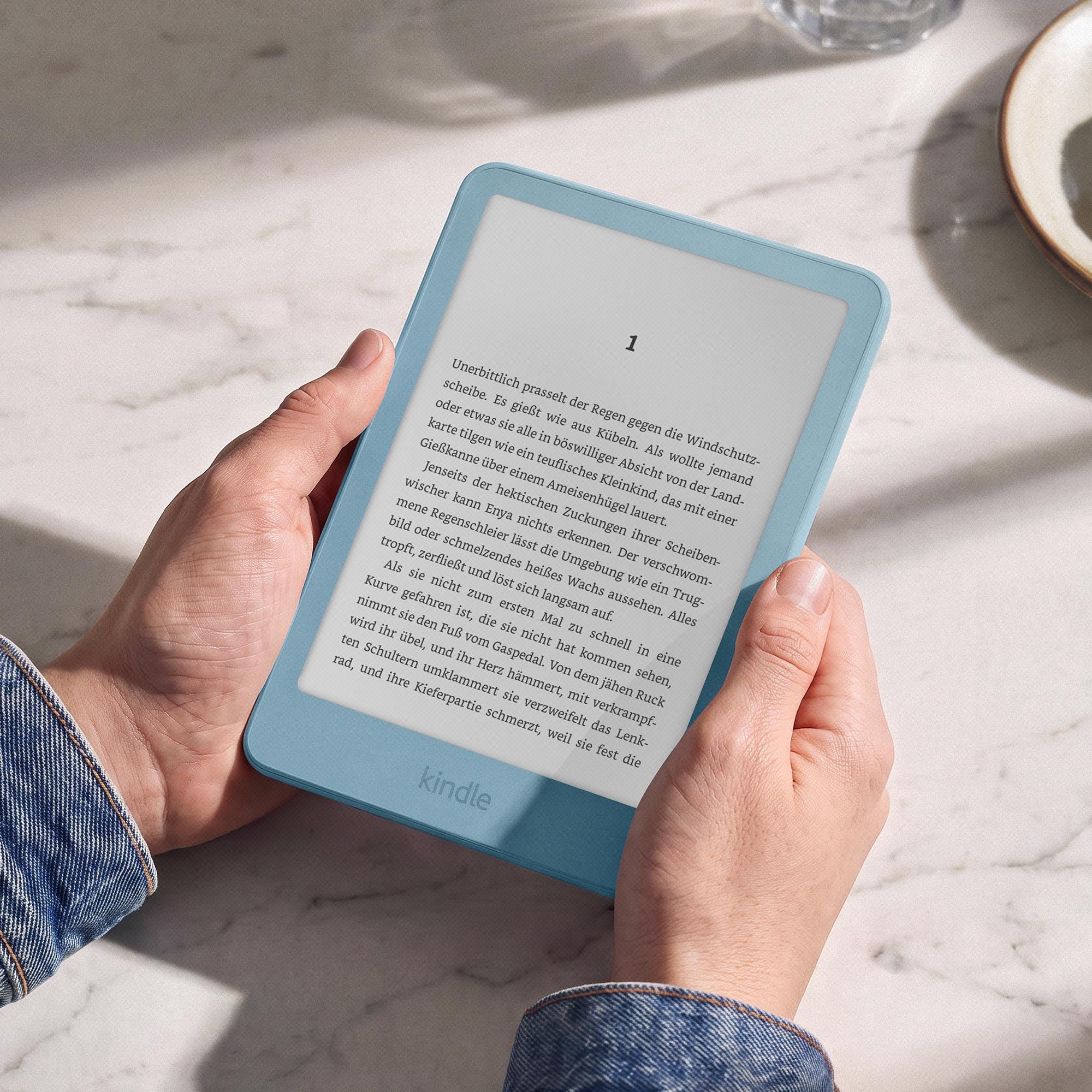 Hände halten einen hellblauen Kindle E-Reader mit deutschem Text auf einem weißen Marmortisch.