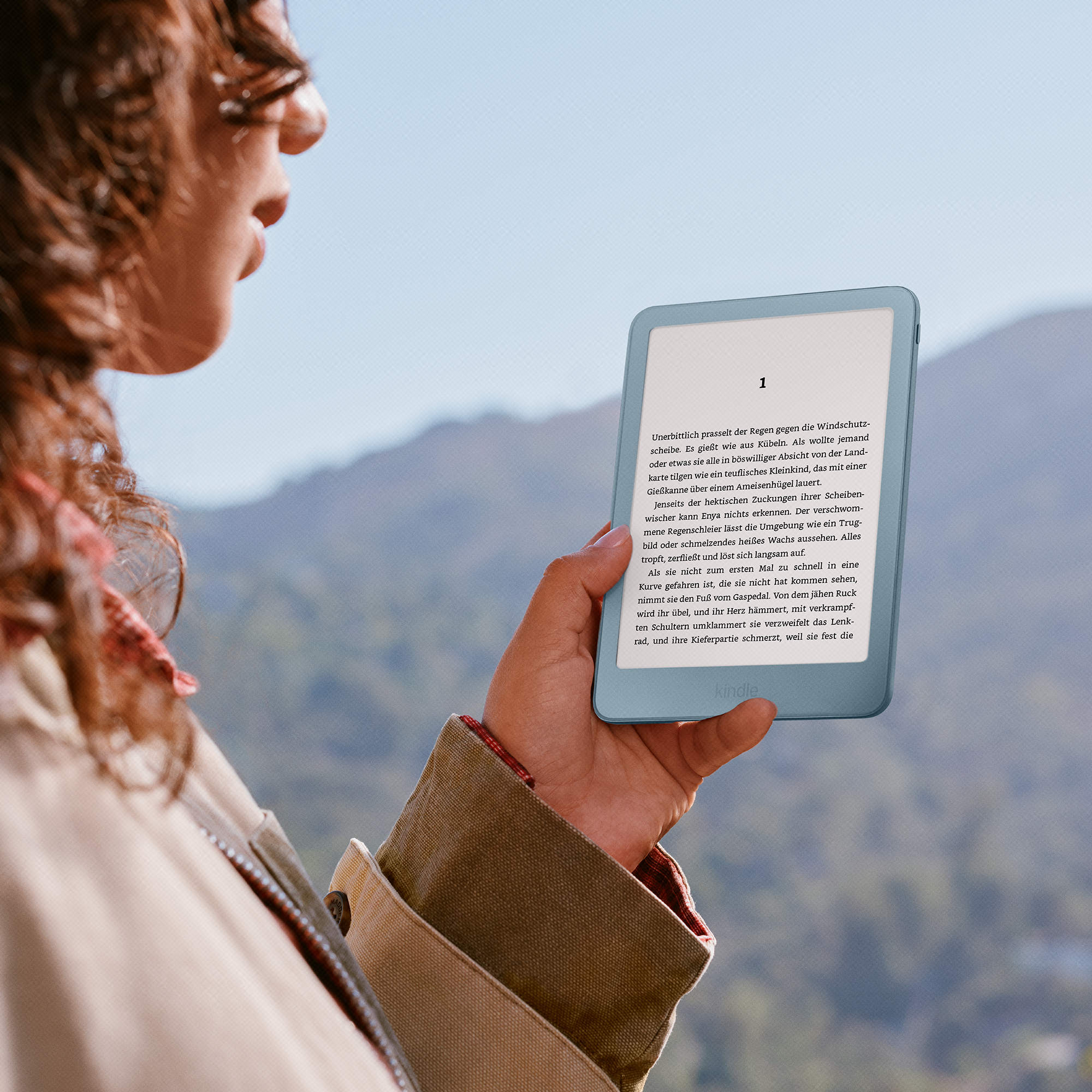 Eine Frau mit lockigem Haar liest ein blaues E-Reader-Gerät im Freien mit Bergen im Hintergrund.