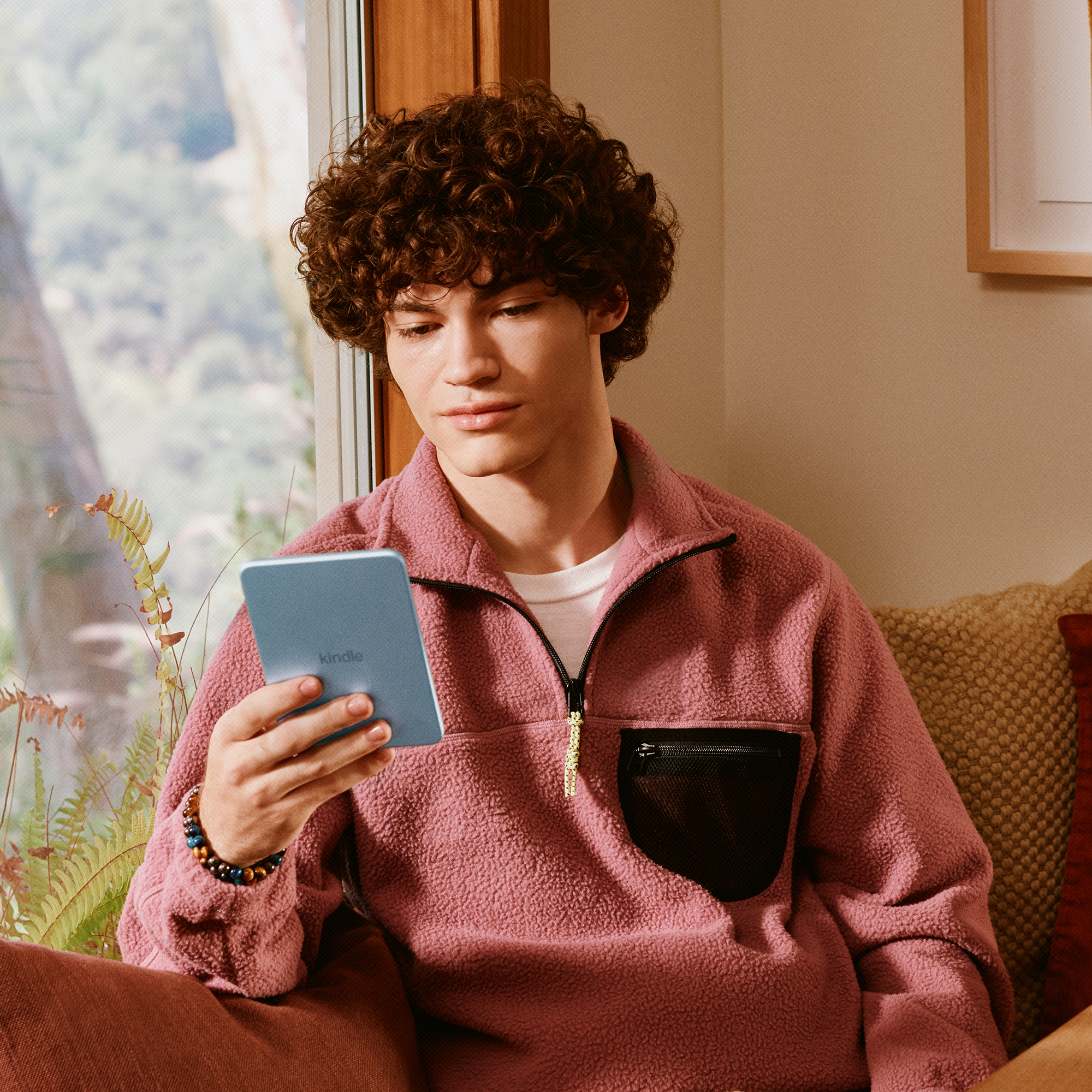 Ein lockiger junger Mann in einem rosa Fleecepullover sitzt drinnen und liest auf einem blauen Kindle.